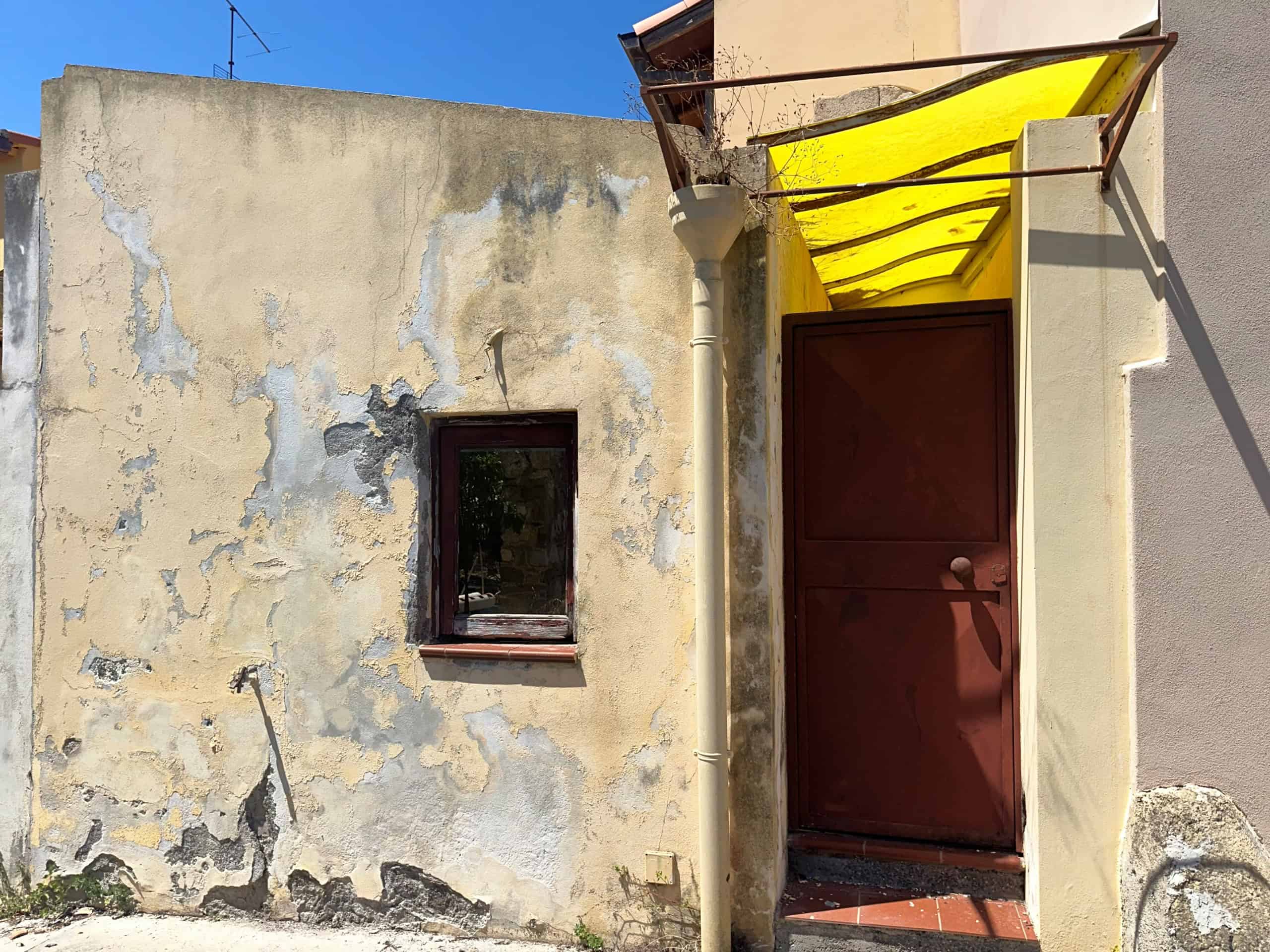 Ingresso di una casa indipendente in Via Trinità 32, Milazzo, con porta rossa e tettoia gialla, pareti esterne con segni di usura.