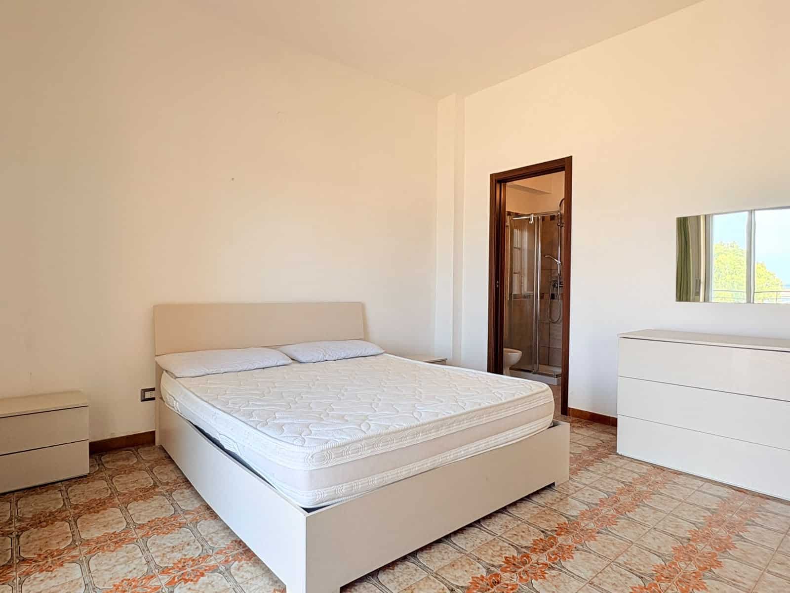 Camera che mostra una camera da letto luminosa con letto matrimoniale, comodino, cassettiera e finestra con vista sul verde.