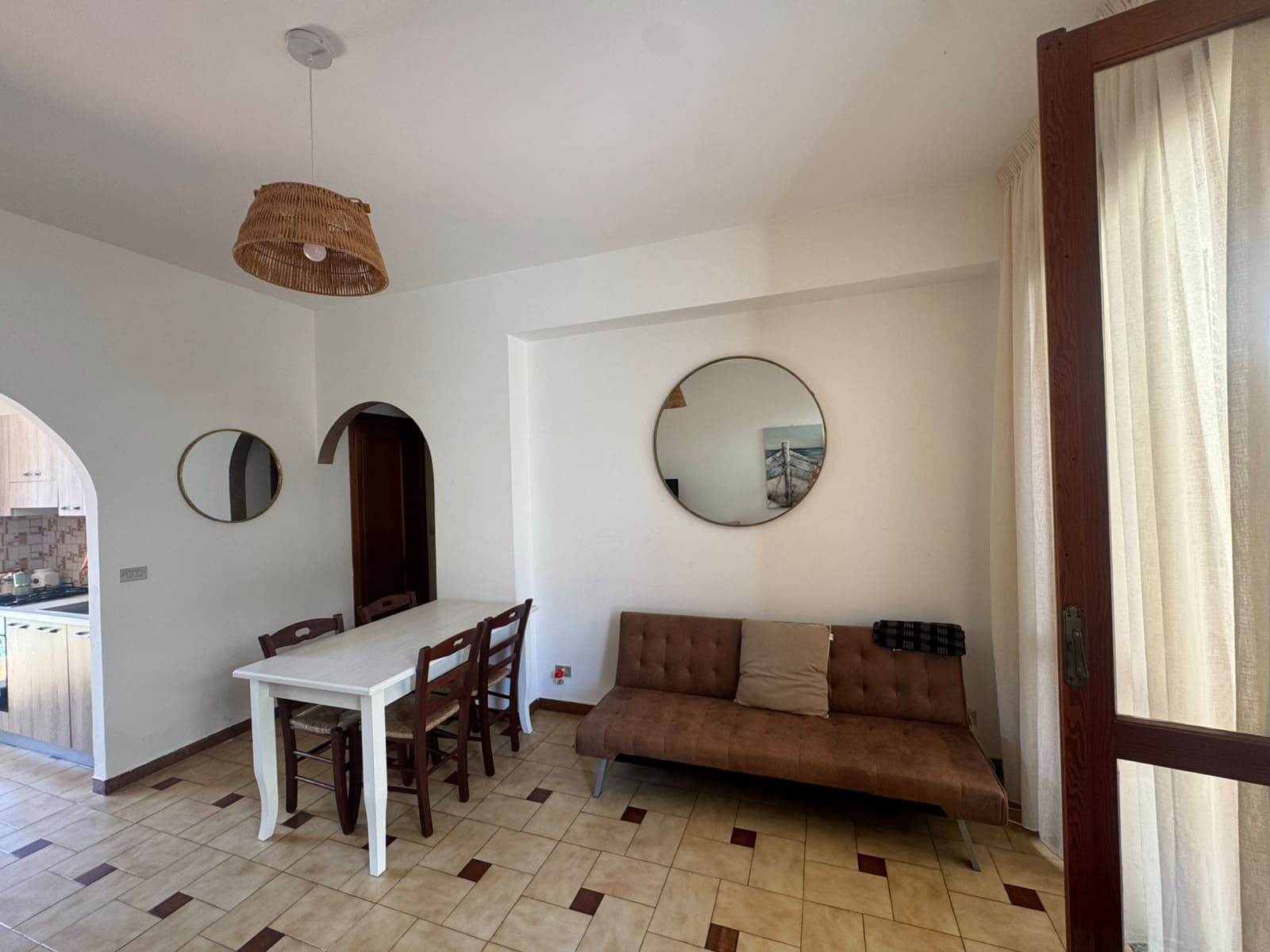 Interno di un soggiorno con divano marrone, tavolo bianco e specchi rotondi sulla parete, illuminato da luce naturale da porta finestra.
