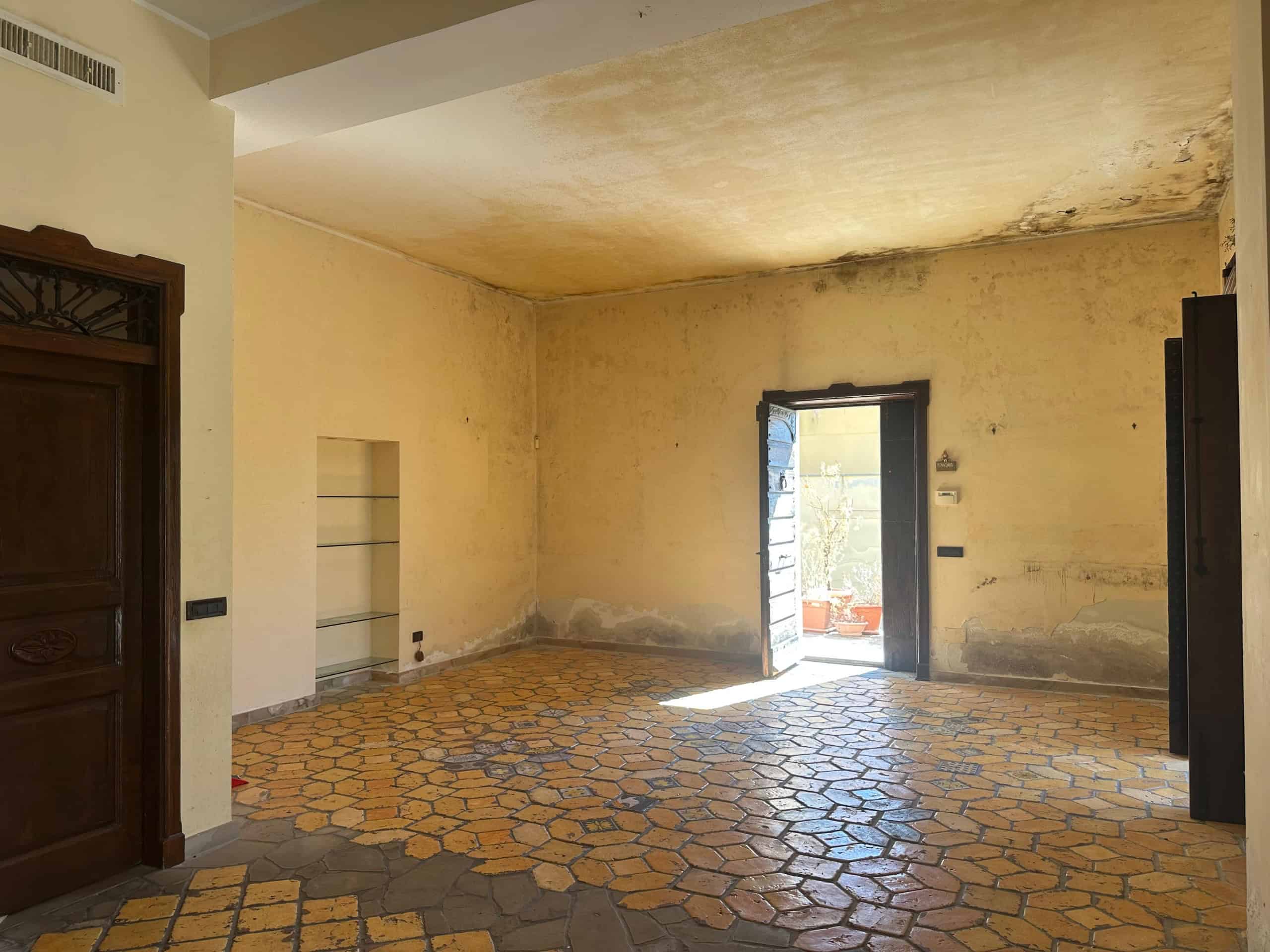 Interno di un appartamento in vendita a Milazzo, con pareti da rinnovare e pavimento in piastrelle colorate, luce naturale proveniente dalla porta aperta.