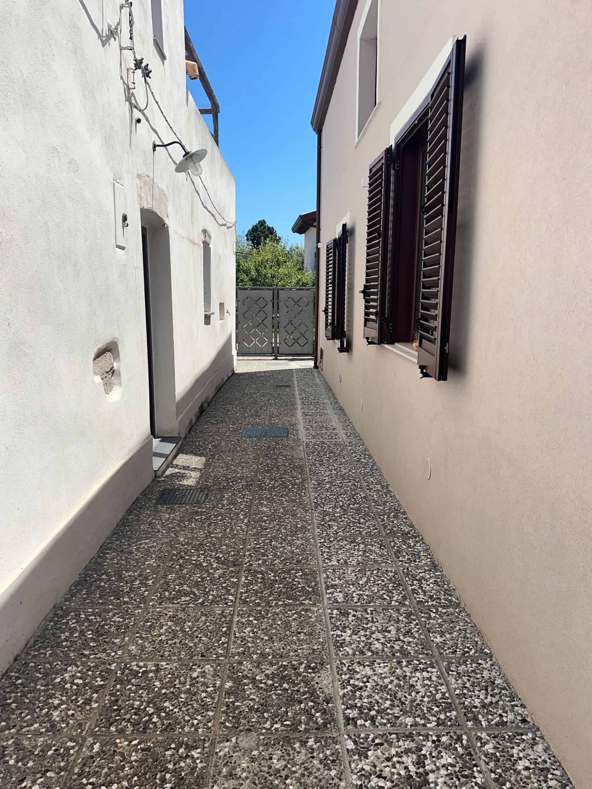 Vicolo tra case in vendita a Milazzo con pavimentazione in pietra e finestre con persiane in legno.