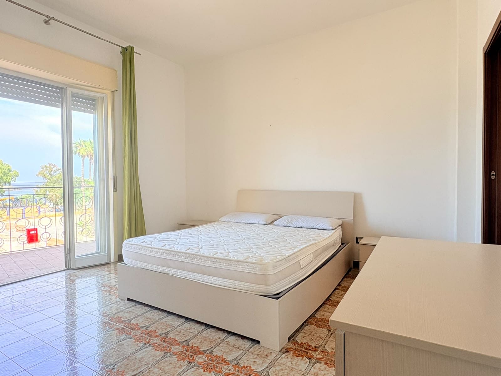 Camera da letto luminosa con balcone vista mare, arredamento semplice e moderno, pavimento in ceramica, ideale per affitti a Milazzo.