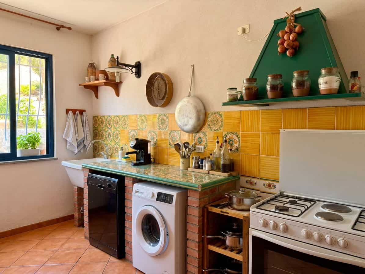 Cucina accogliente con lavatrice, forno e scaffali decorativi, illuminata dalla luce naturale dalla finestra.