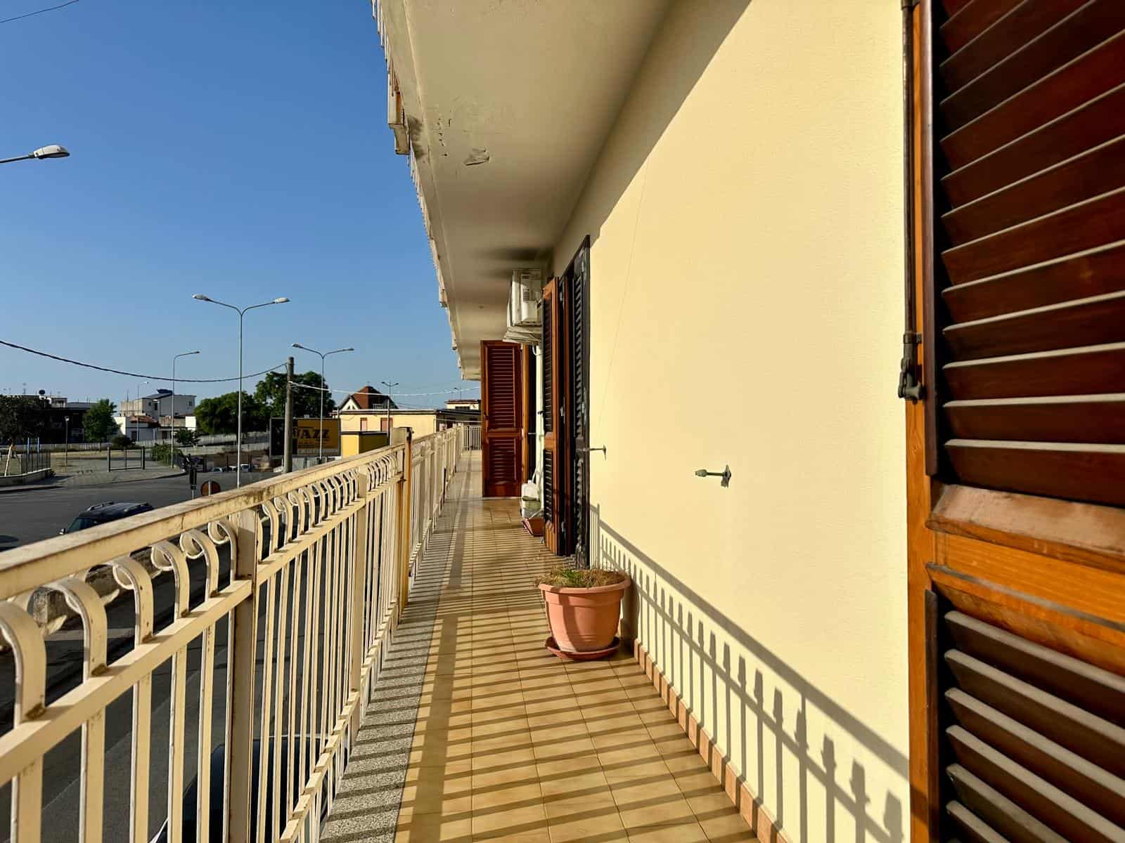 Balcone di un appartamento con ringhiera in ferro battuto e piante in vaso, vista sulla strada e il cielo azzurro.