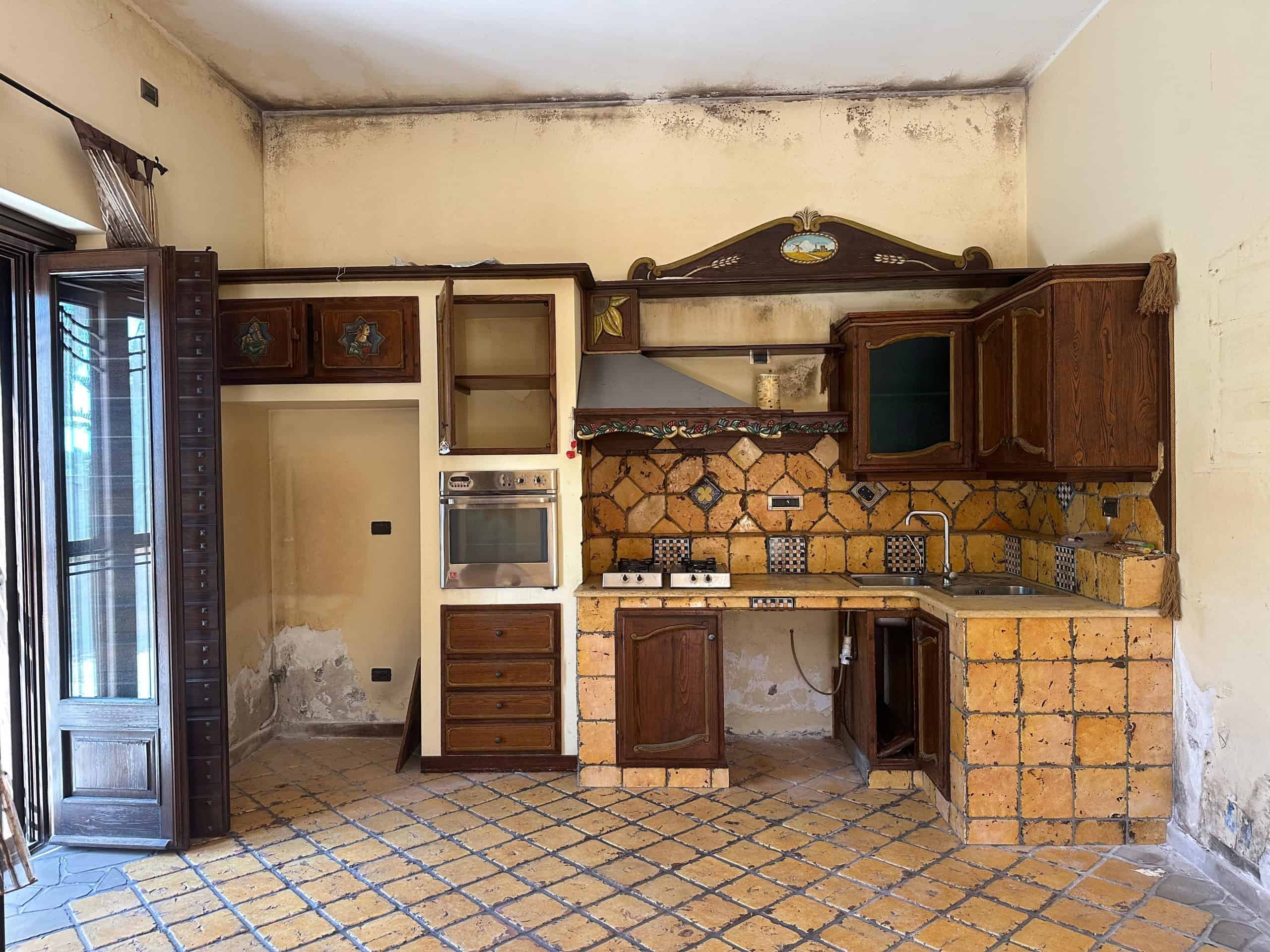 Cucina rustica in condizioni di abbandono con mobili in legno e pareti danneggiate, pavimento in piastrelle e soffitto con muffa.