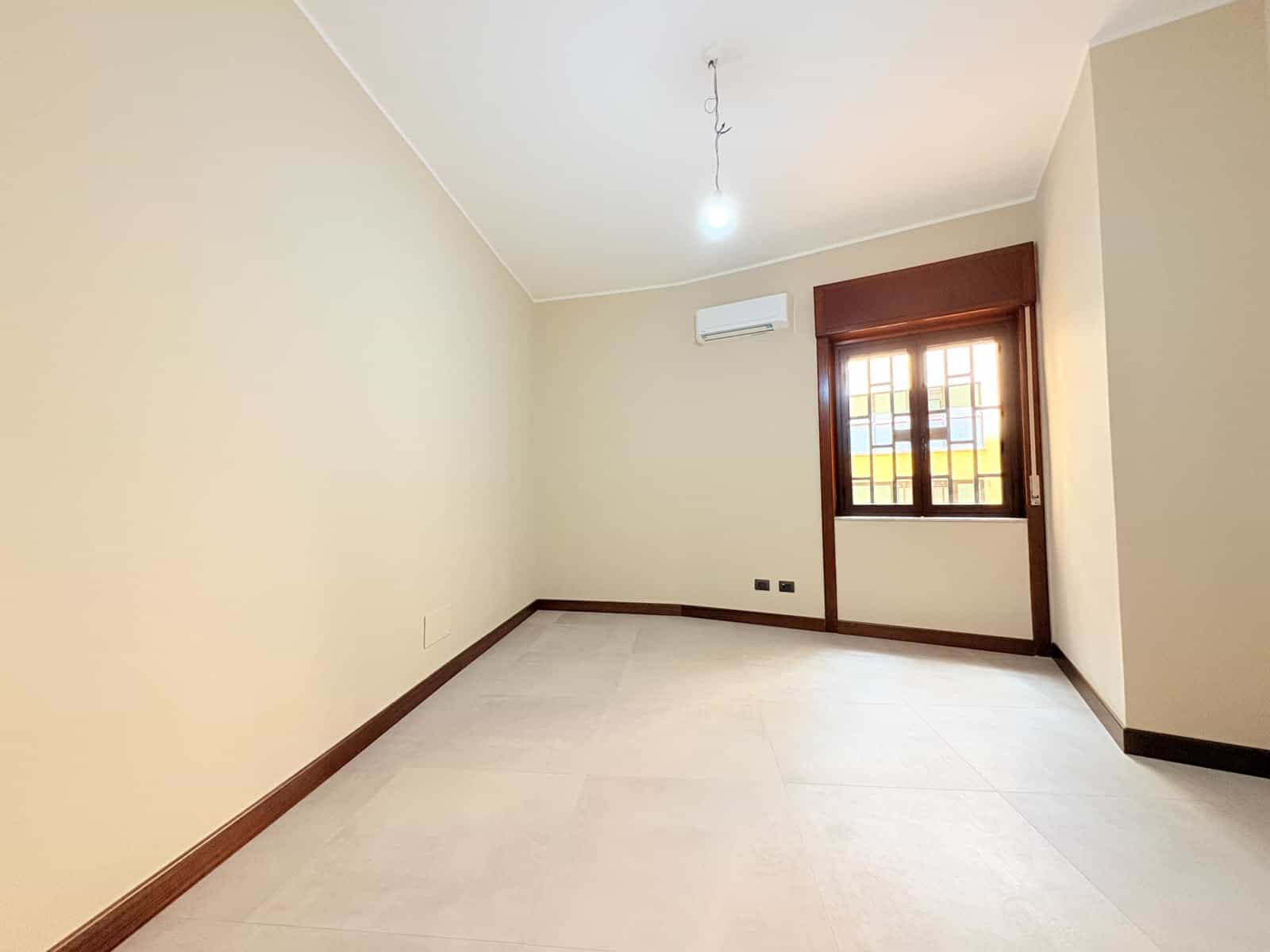 Stanza luminosa con pareti beige, finestra con infissi in legno e aria condizionata.
