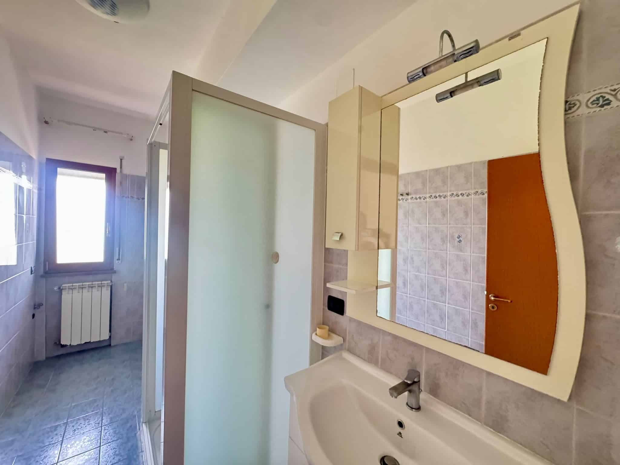 Bagno con lavabo, specchio e doccia, finestra luminosa, pavimento e pareti in piastrelle.