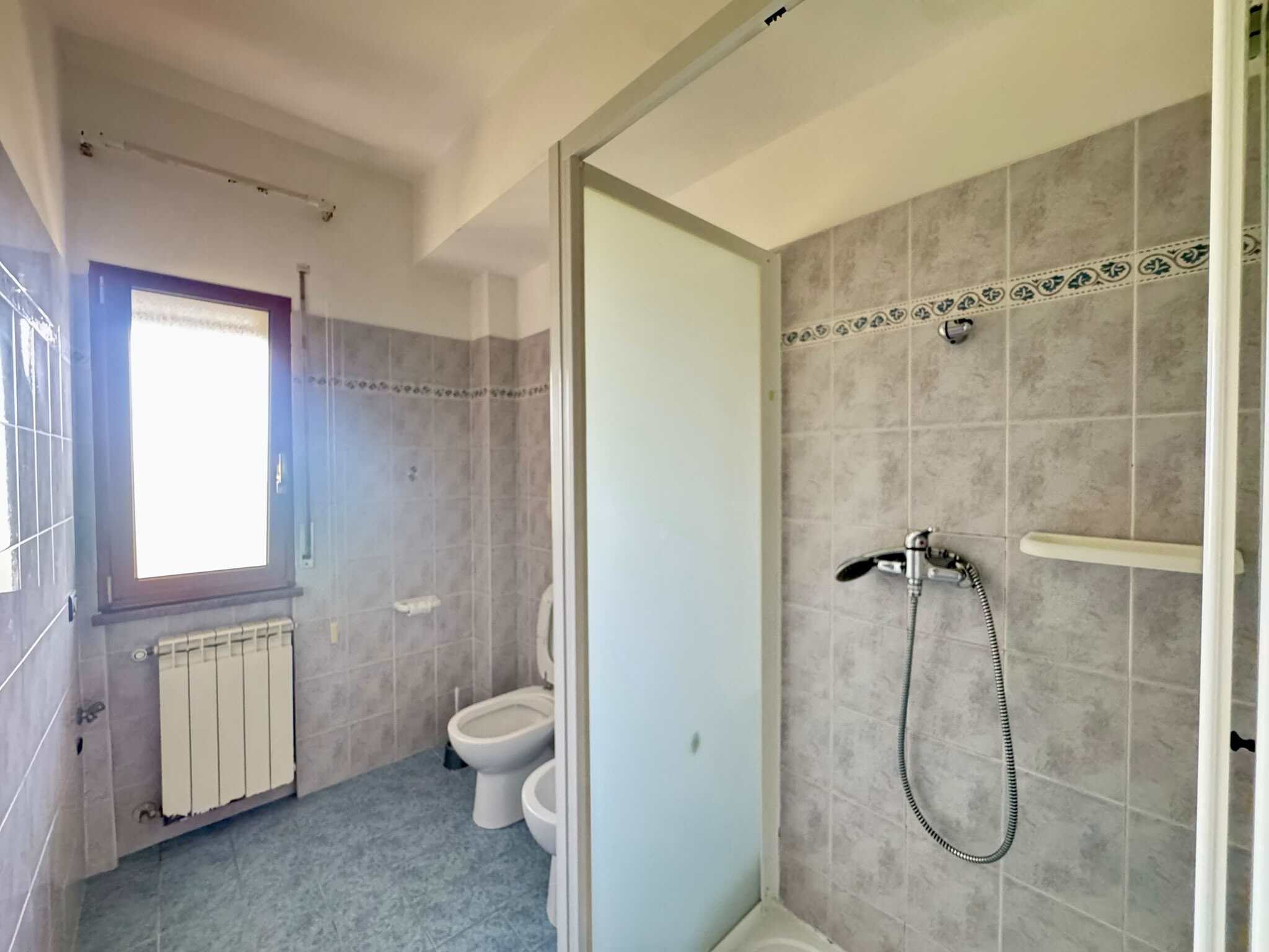 Bagno con doccia, WC e finestra, pareti rivestite in piastrelle grigie, ambiente luminoso e funzionale.