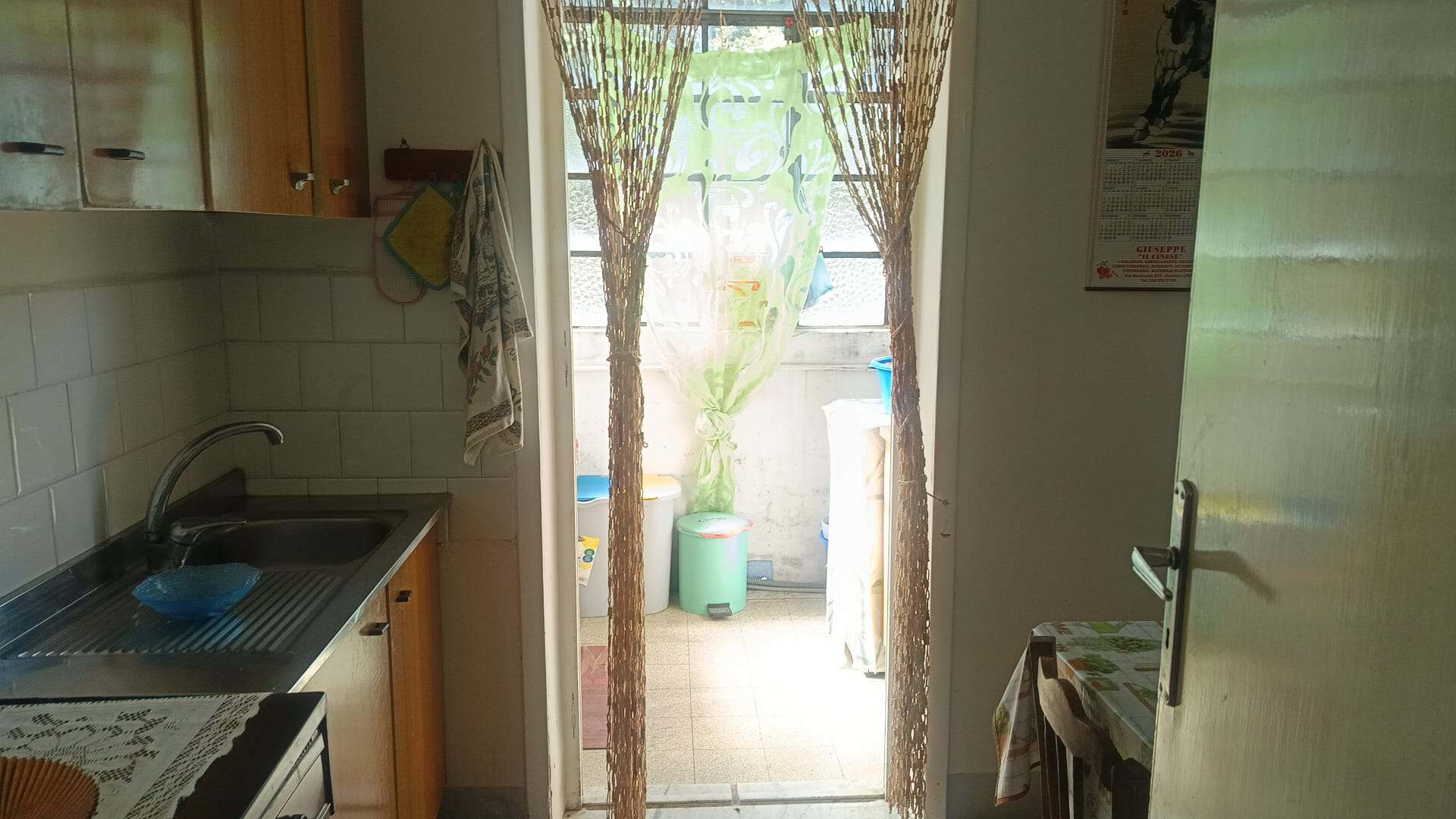 Vista dell'ingresso di un appartamento con porta aperta, cucina e accesso a un balcone esterno con piante e oggetti vari.
