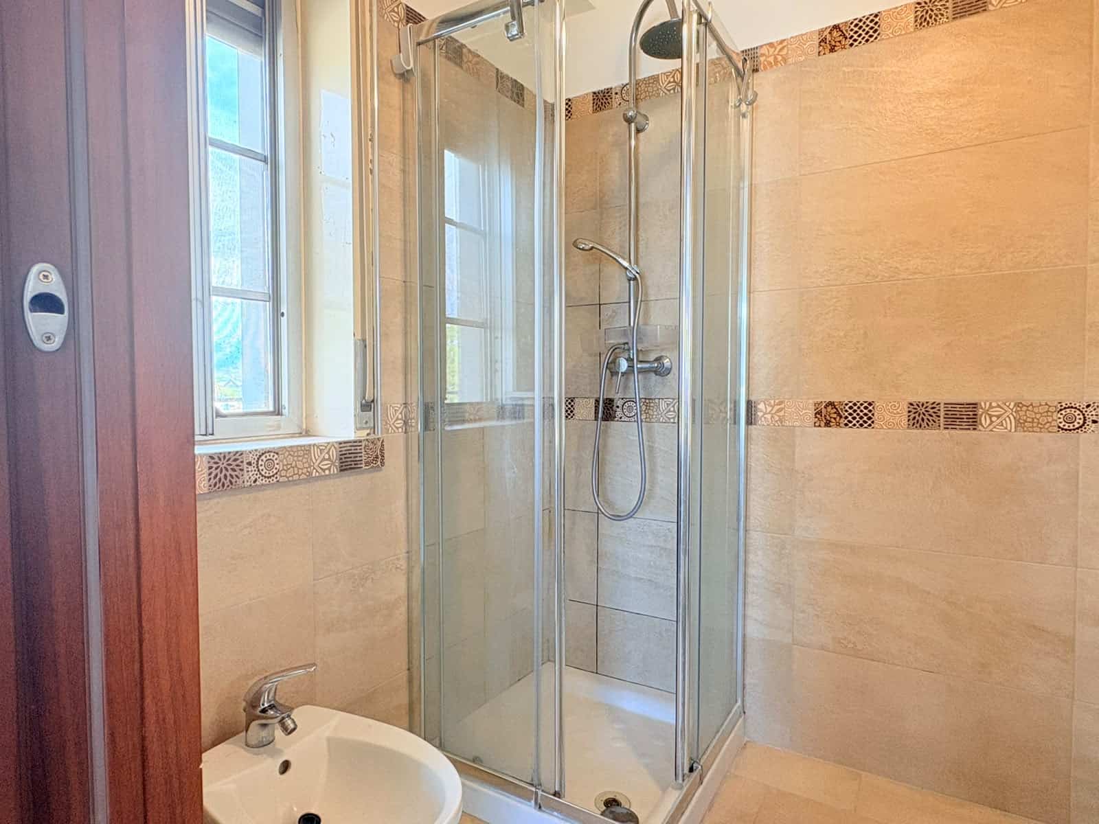 Doccia moderna in bagno con pareti in piastrelle beige e finestra.