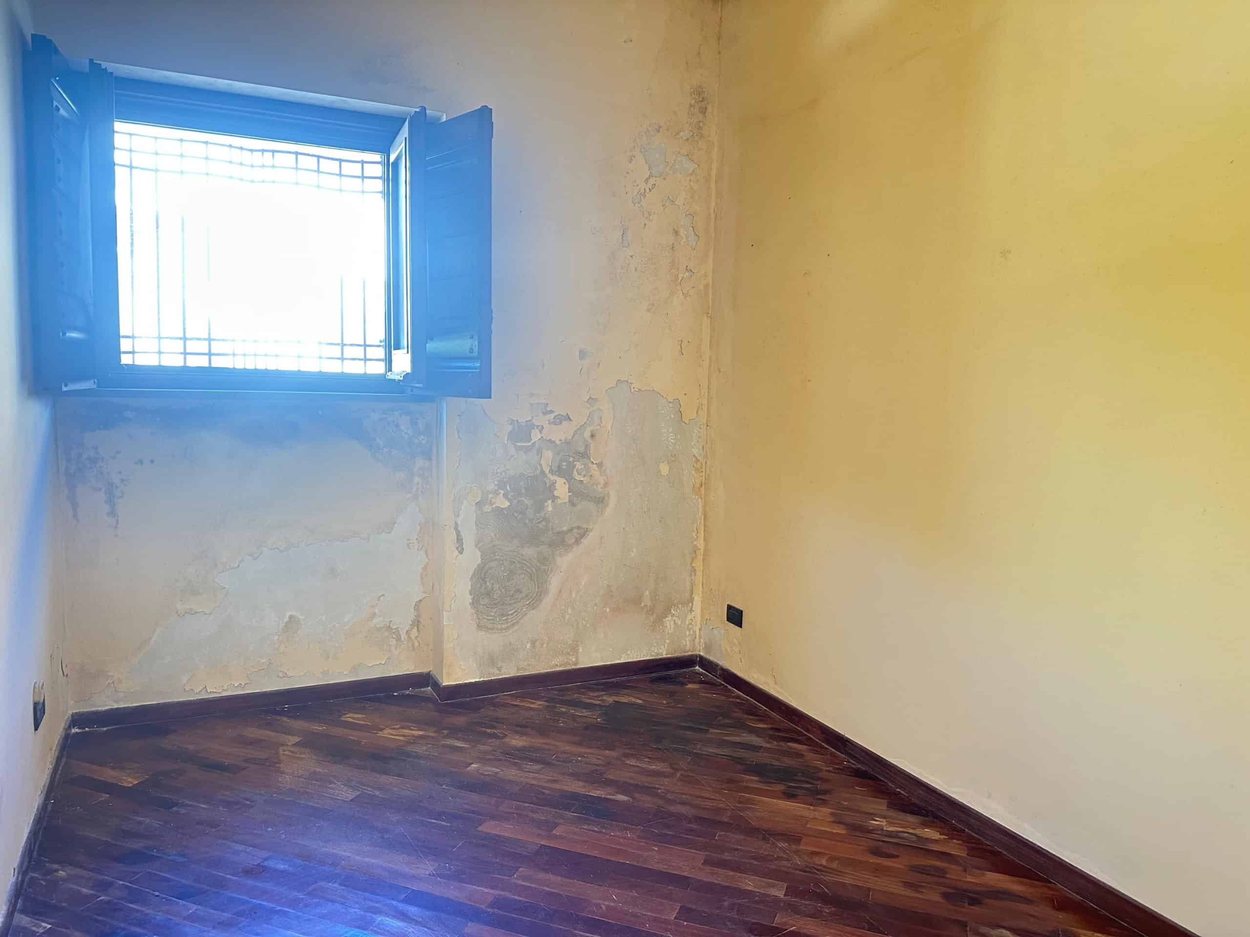 Stanza vuota con pareti usurate e finestra con grate in una proprietà in vendita a Milazzo.