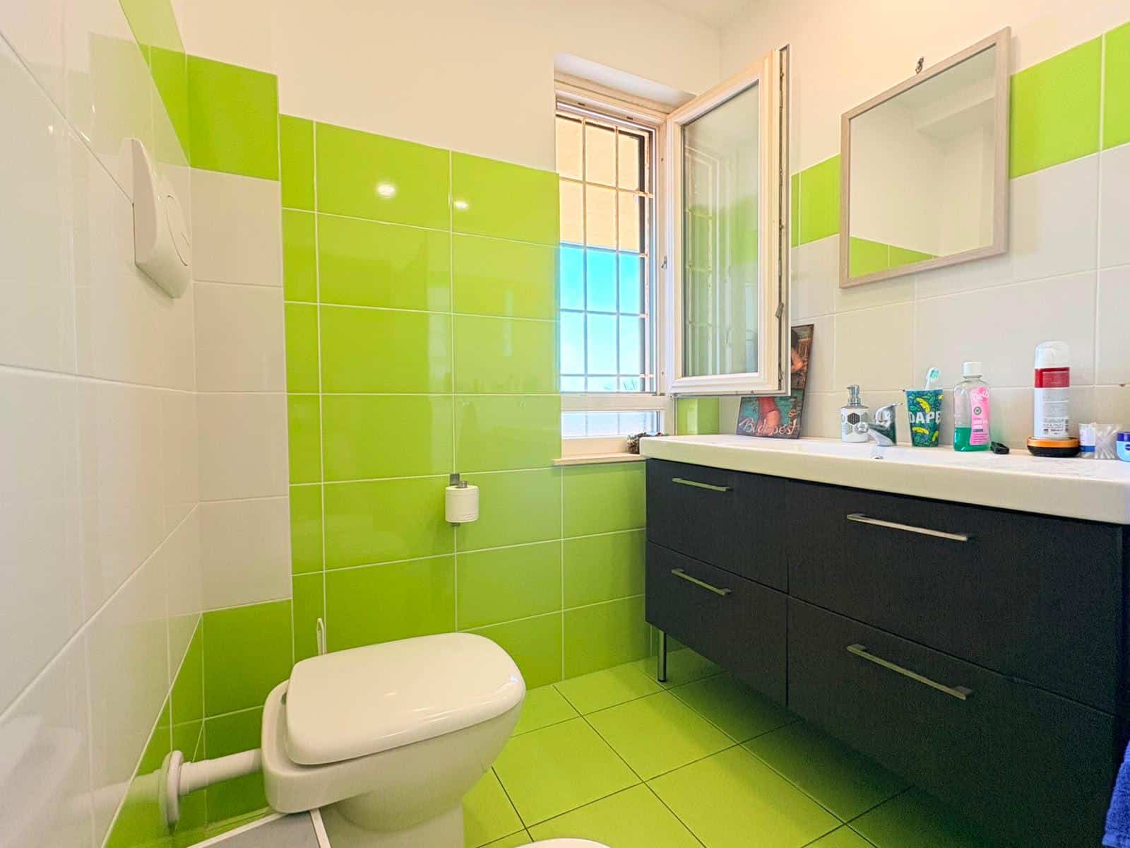 Bagno moderno con pareti verdi e bianco, lavabo e specchio, finestra aperta, in un appartamento in vendita a Milazzo.