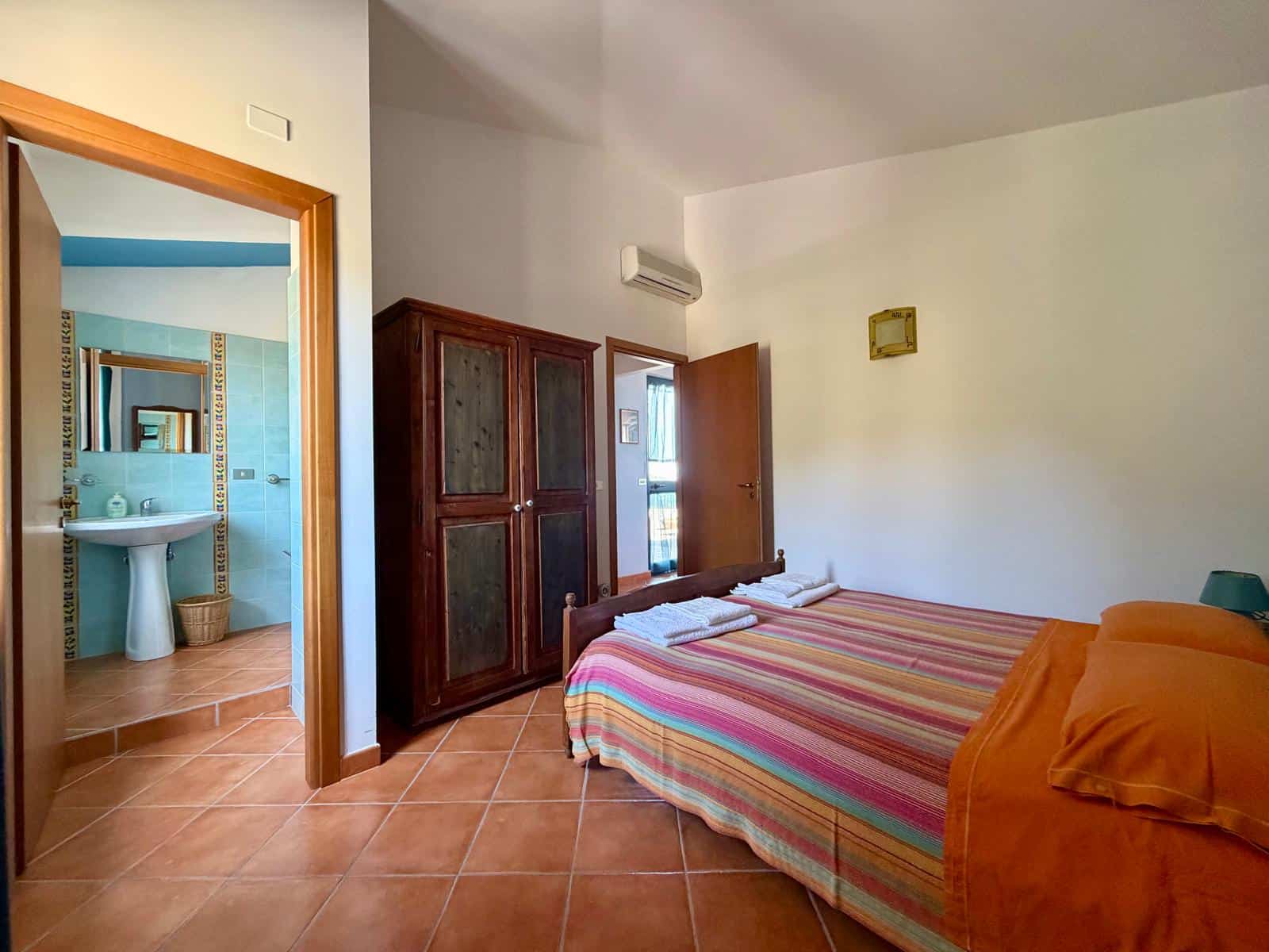 Camera da letto con letto matrimoniale, armadio in legno e bagno visibile dalla porta aperta, ambiente luminoso e accogliente.
