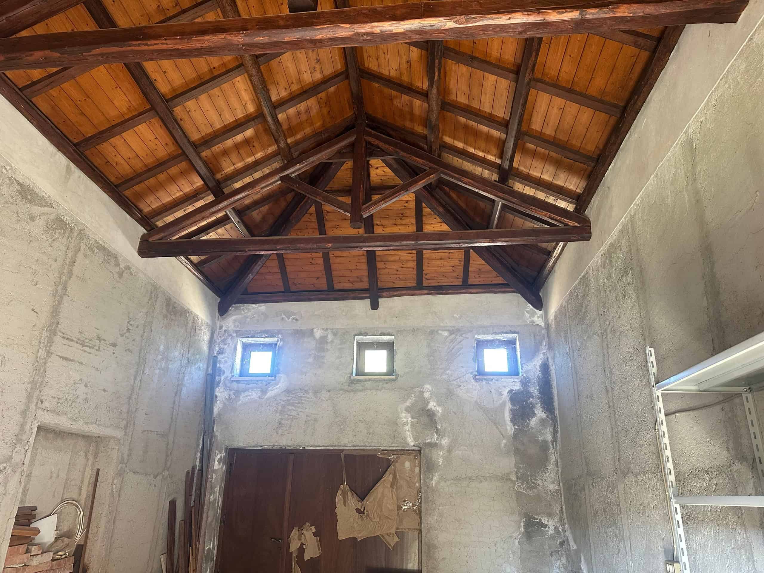 Interno di una stanza con soffitto in legno e pareti in cemento non rifinite, con tre piccole finestre.