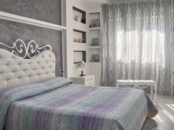 Interno di una camera da letto moderna con letto imbottito, tende leggere e scaffali decorativi, in un appartamento in vendita a Messina.