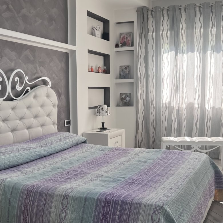 Interno di una camera da letto moderna con letto imbottito, tende leggere e scaffali decorativi, in un appartamento in vendita a Messina.