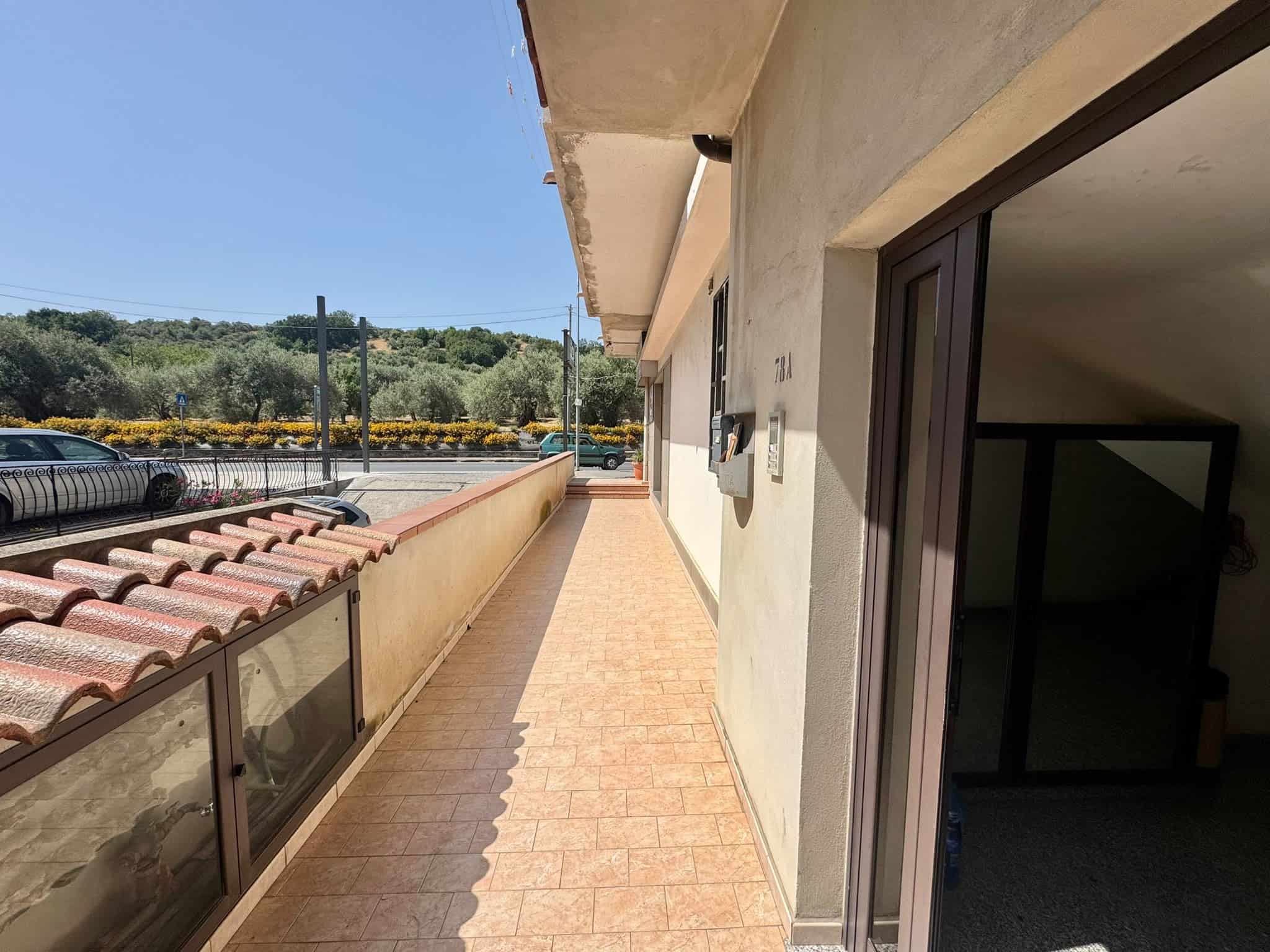 Balcone di un appartamento con vista su un paesaggio verde e cielo azzurro, con pavimentazione in piastrelle e pareti beige.