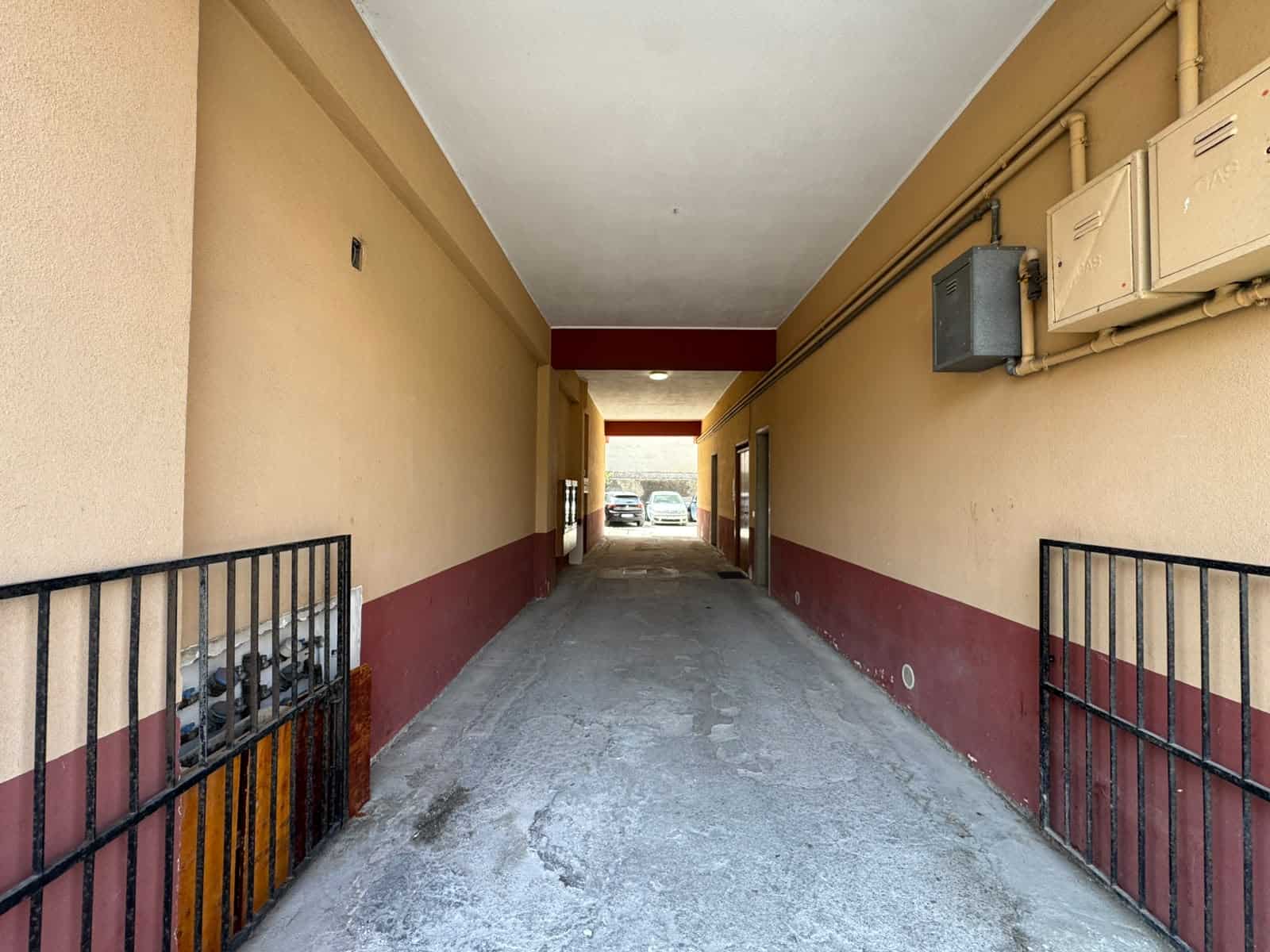 Passaggio coperto in un condominio con pareti beige e rosse, cancelletti in ferro e cablaggi elettrici visibili, parcheggio all'aperto in fondo.