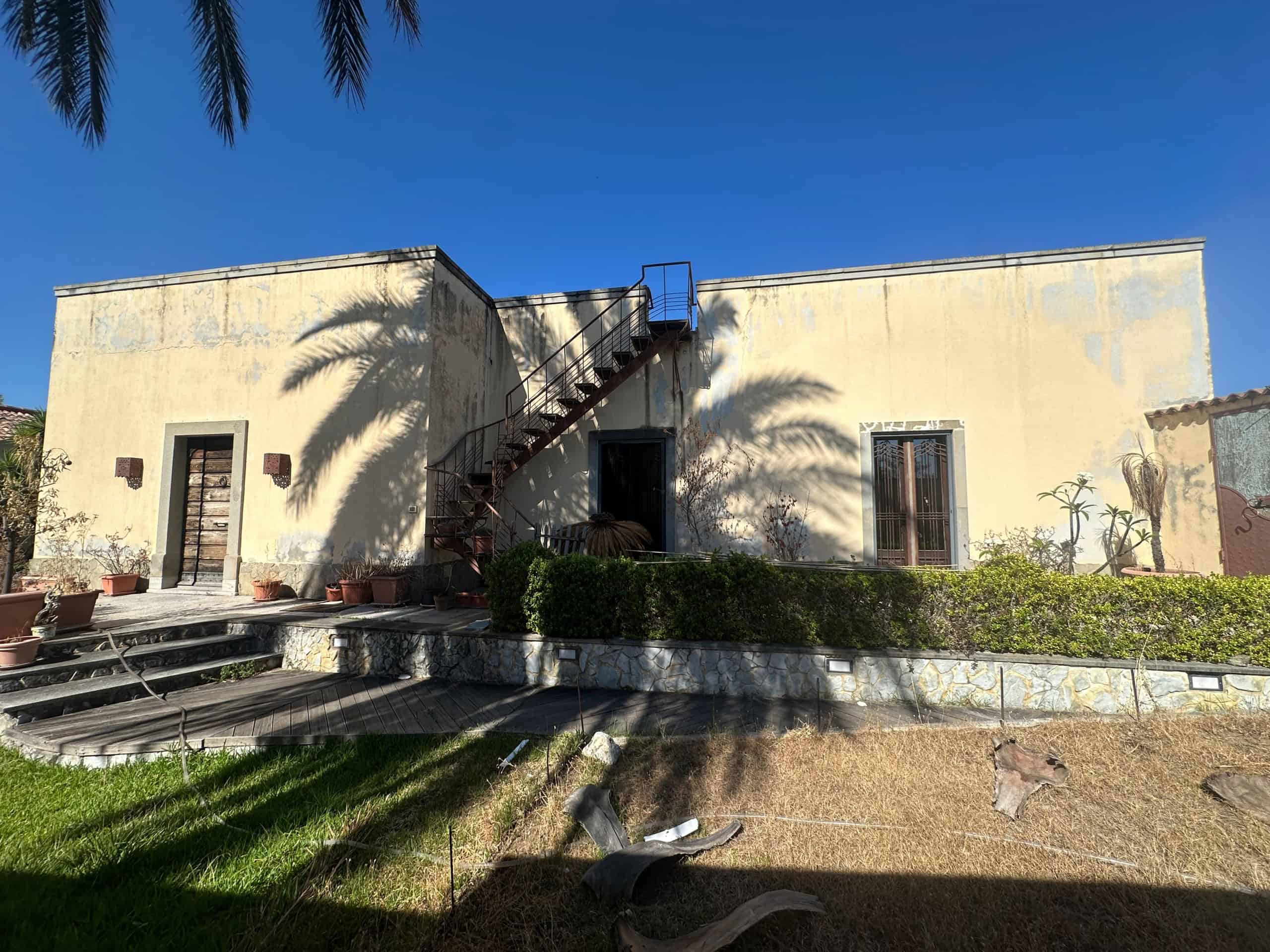 Villa in vendita a Milazzo con facciata bianca, scale esterne e giardino ben curato sotto un cielo sereno.