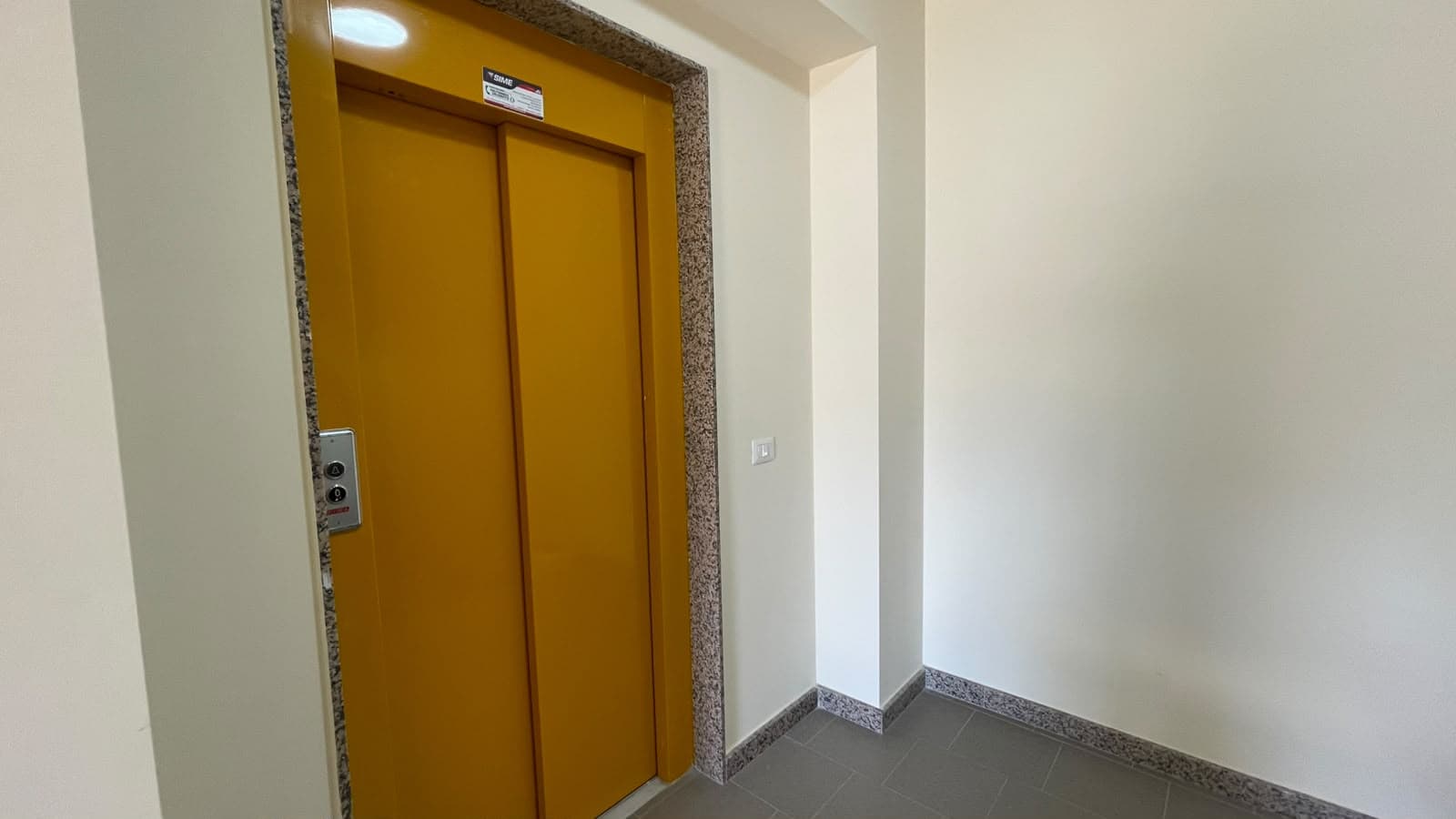 Ascensore giallo in edificio residenziale a Spadafora, ME, con pareti bianche e pavimento in piastrelle grigie.
