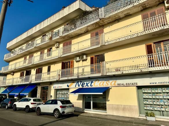 Edificio commerciale con negozio NextCasa e parcheggio di fronte, in zona urbana.