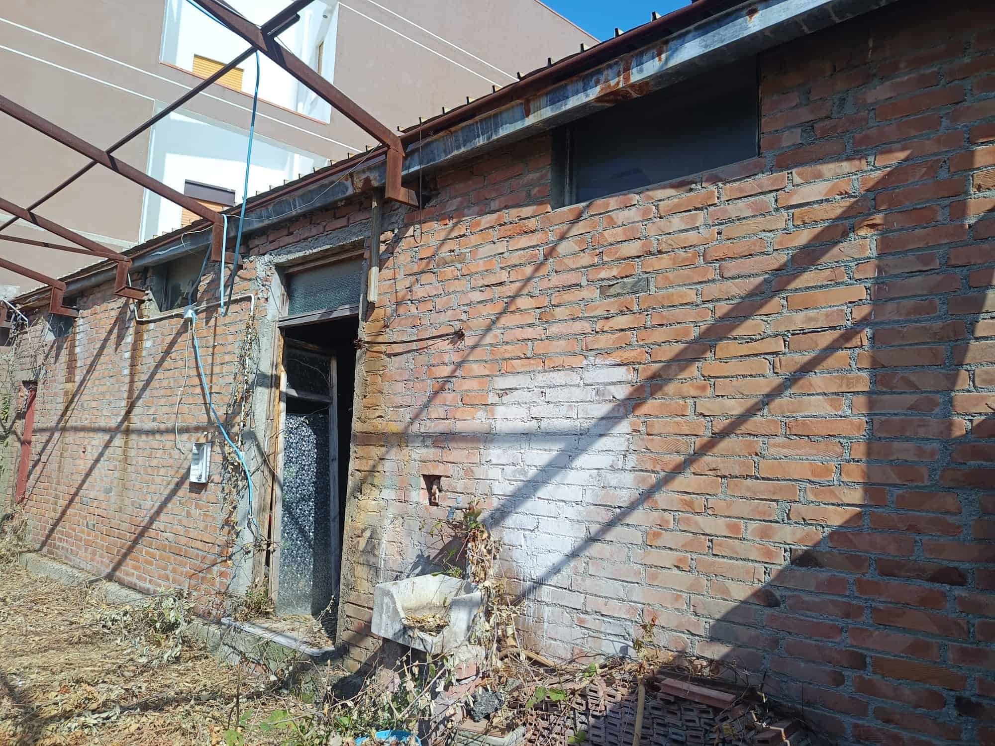 Parete esterna di un edificio in mattoni con porta e struttura metallica sopra, ombreggiata da linee di luce e ombra.