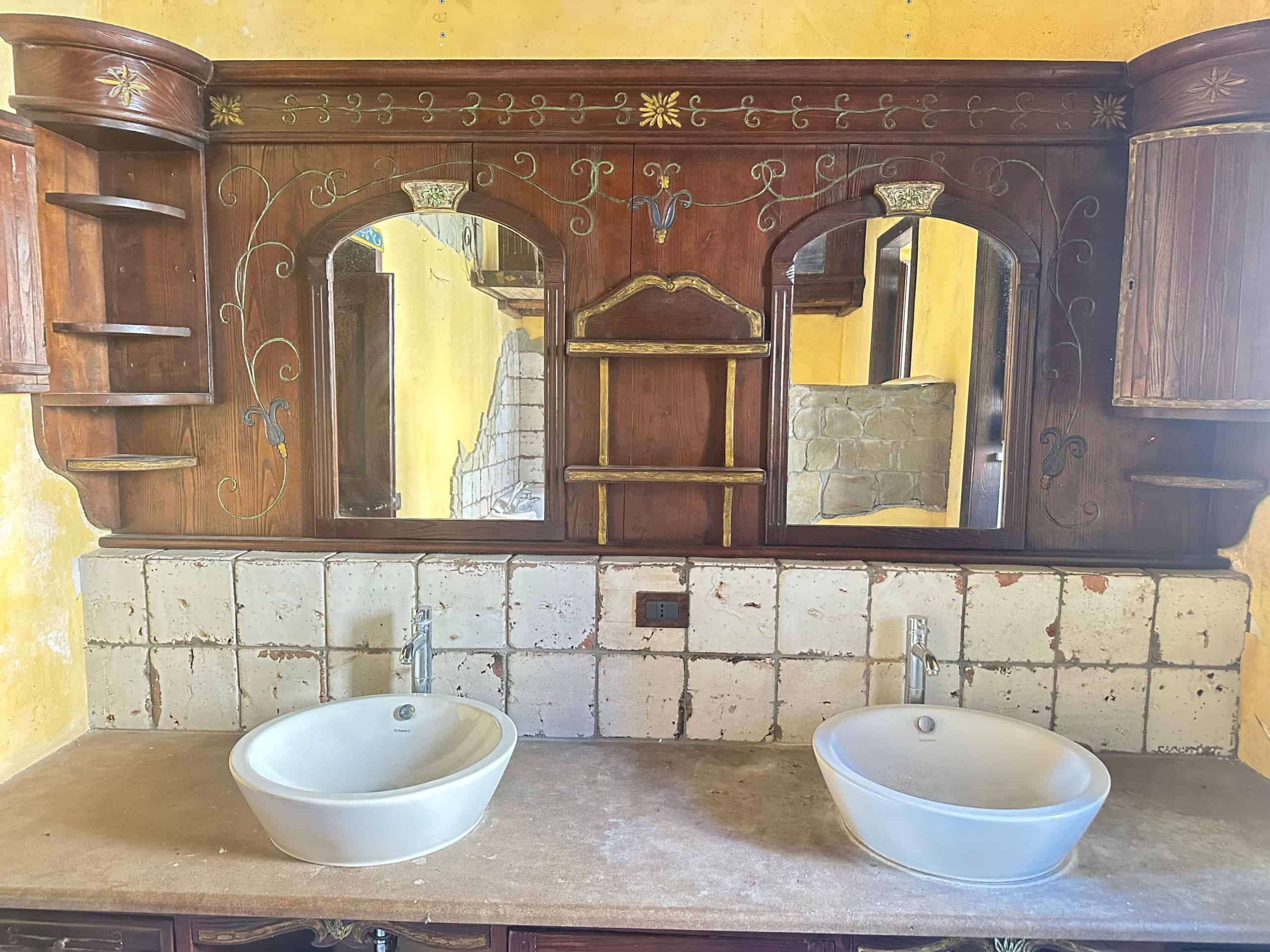 Antico mobile bagno in legno con due lavabi bianchi e specchi, stile rustico, in un ambiente con pareti in pietra e dettagli decorativi in oro.