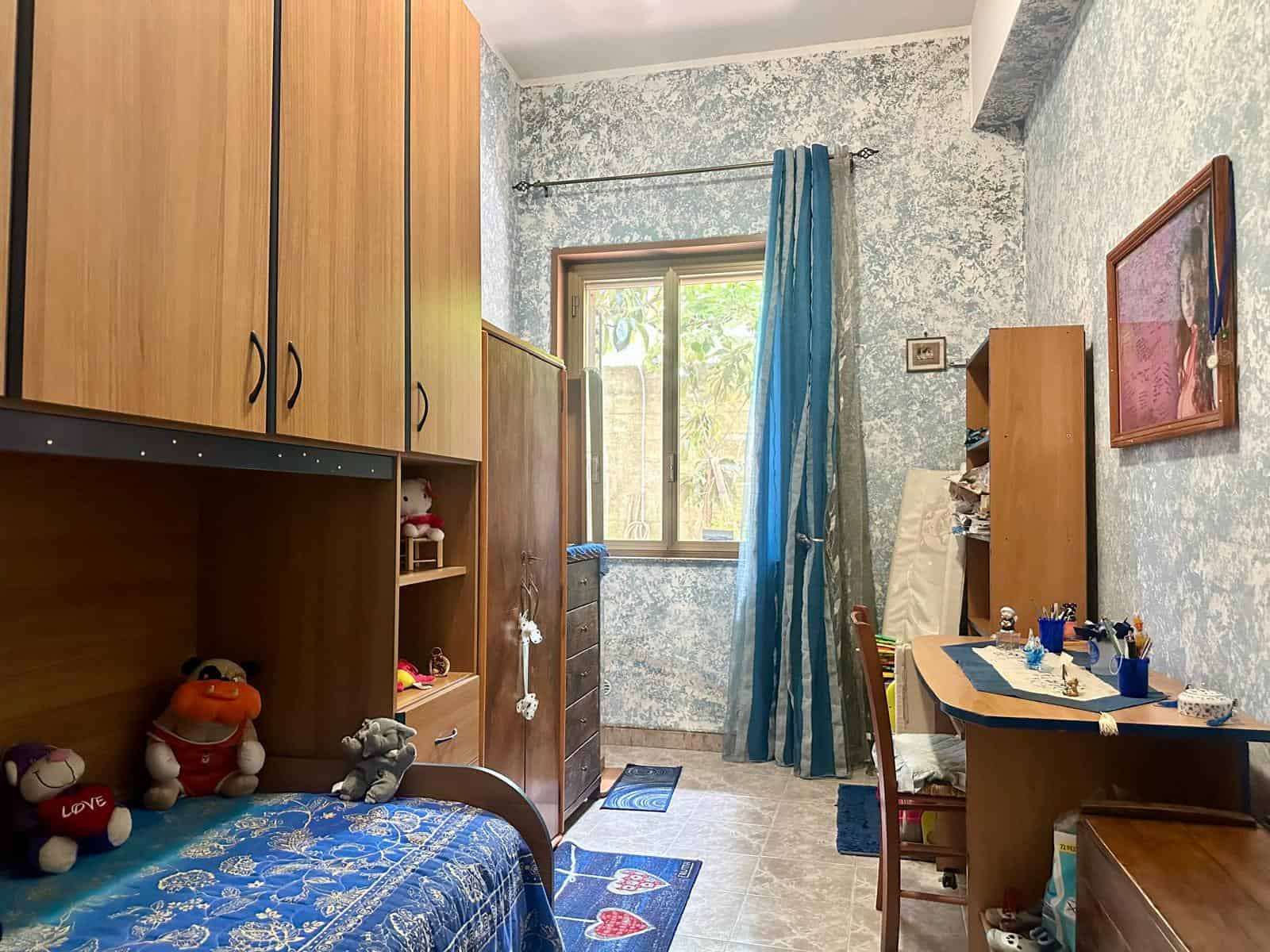Camera da letto con armadio, scrivania e finestra con tende blu, pareti con decorazioni e giochi sul letto.
