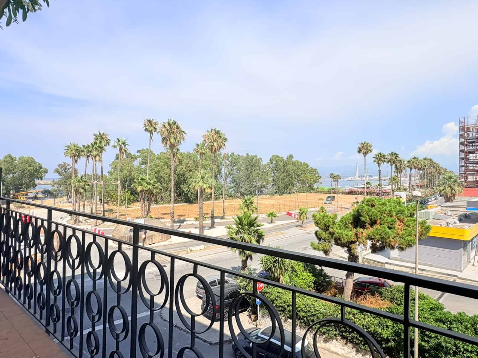 Vista panoramica di un'area urbana con palme, parcheggi e edifici, vista dal balcone di un appartamento in affitto a Milazzo.