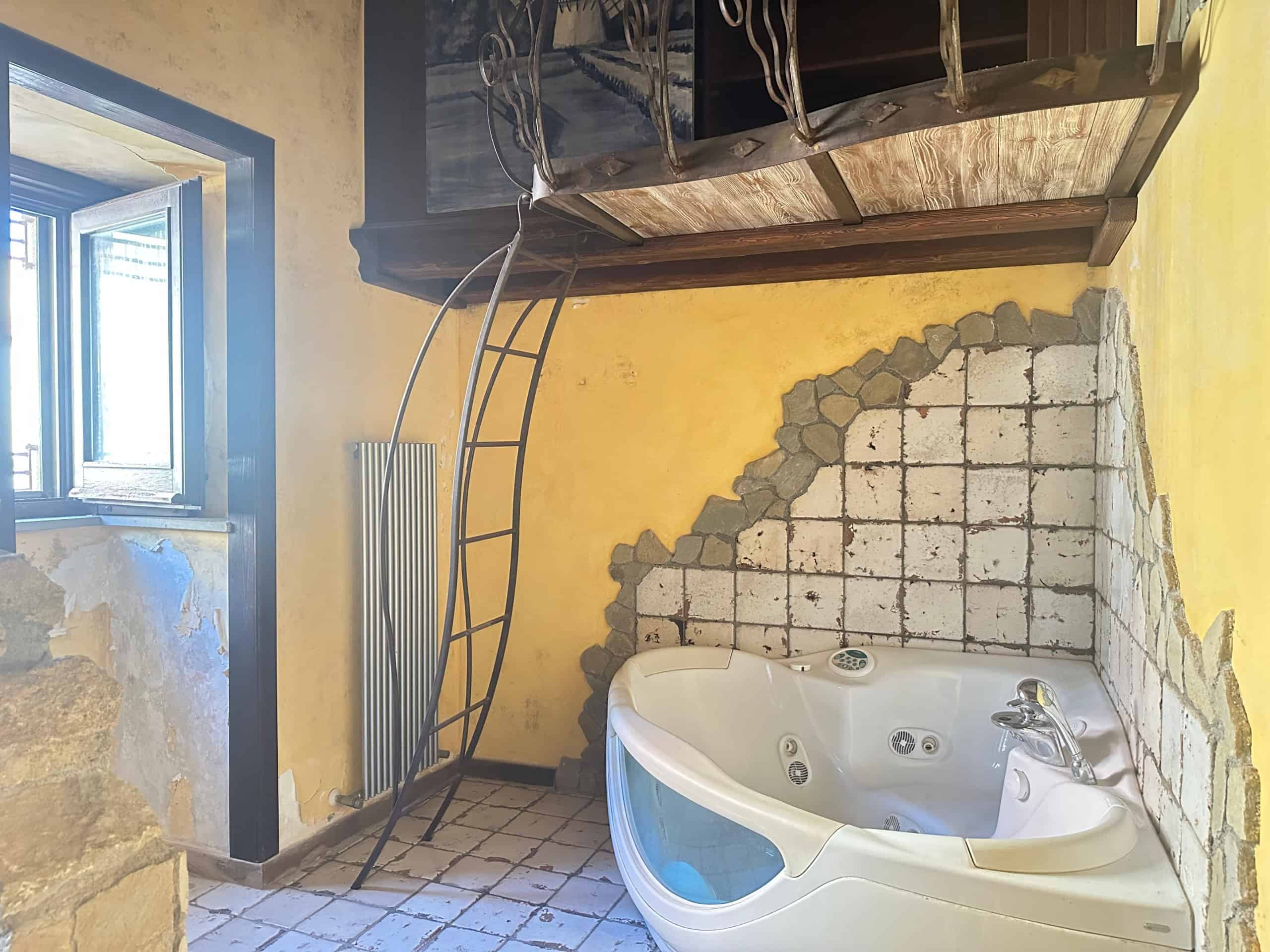 Interno di una stanza con vasca idromassaggio, pareti in pietra e soffitto in legno, illuminata da una finestra aperta.