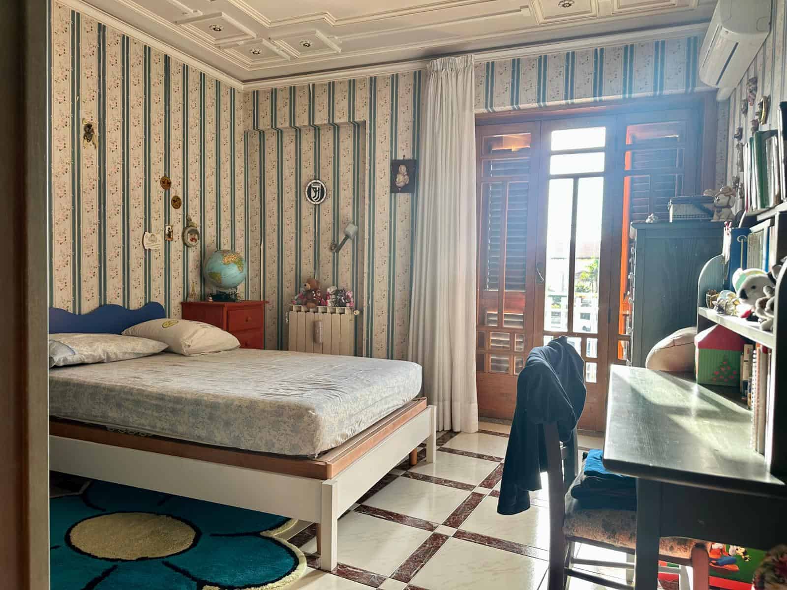 Camera da letto luminosa con pareti a righe blu e beige, grande finestra con persiane in legno e balcone, letto matrimoniale, scrivania con sedia, decorazioni e oggetti vari.