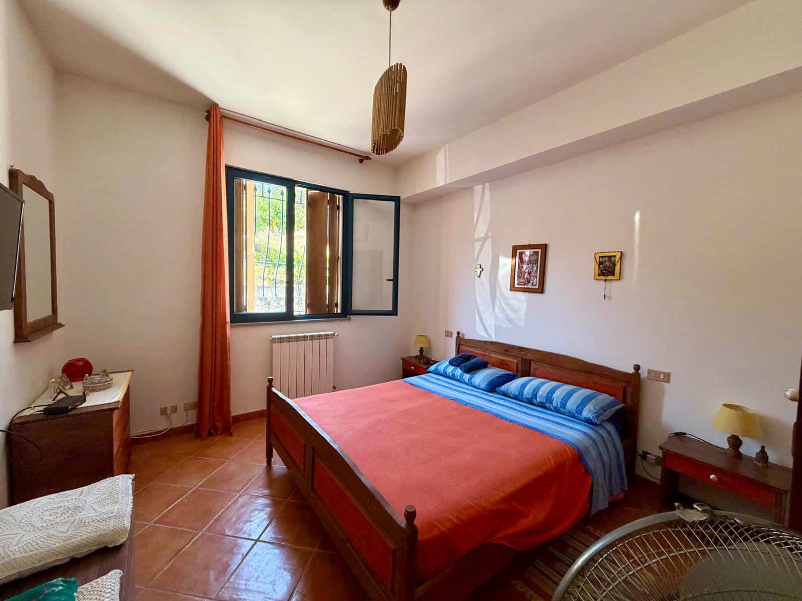 Camera da letto luminosa con letto in legno, lenzuola blu e arancioni, finestra aperta con tende arancioni, arredamento semplice e pareti bianche.