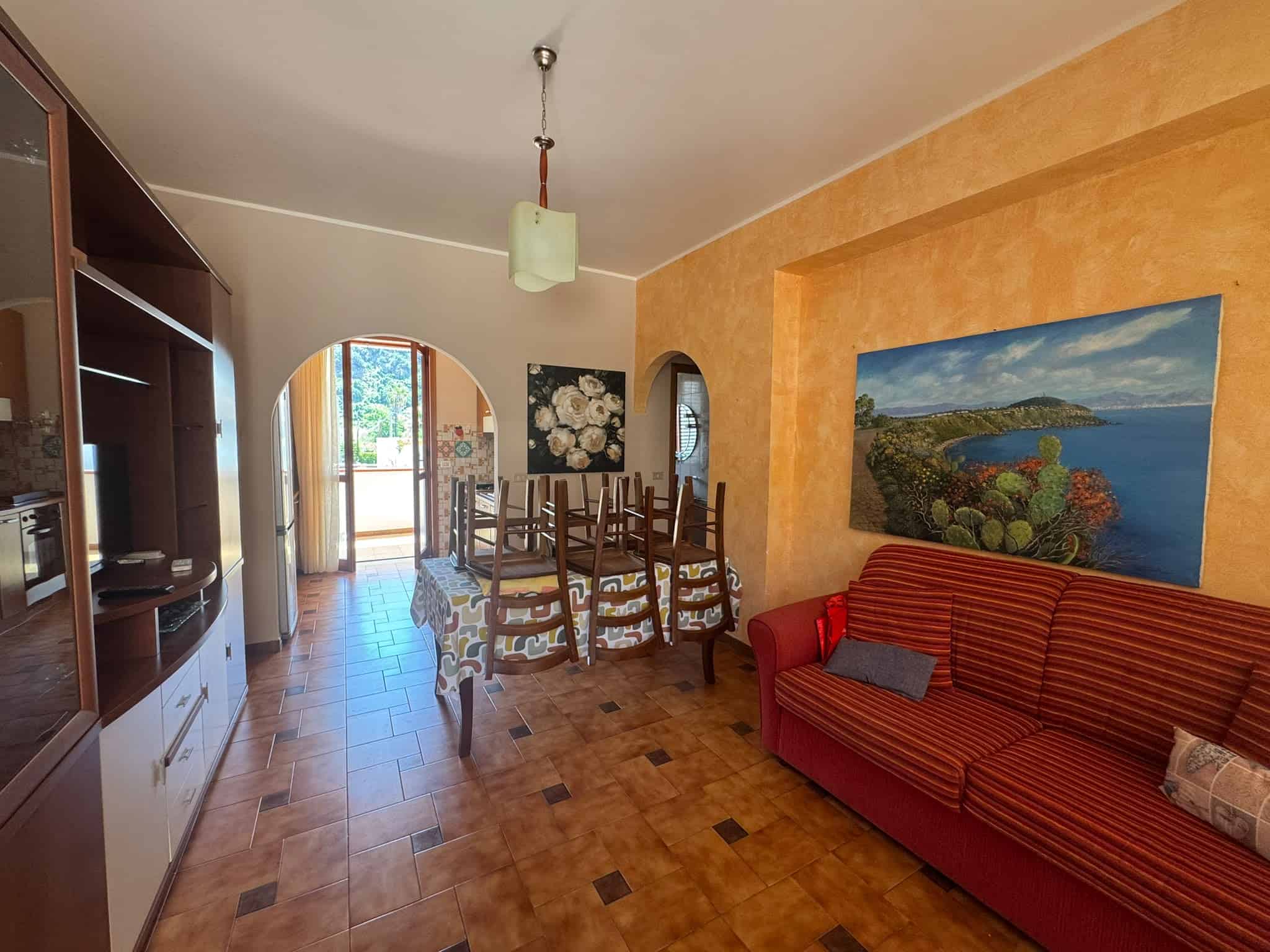 Interno di una villa in vendita a Milazzo, soggiorno con pareti arancioni, divano rosso e sala da pranzo con tavolo e sedie in legno.