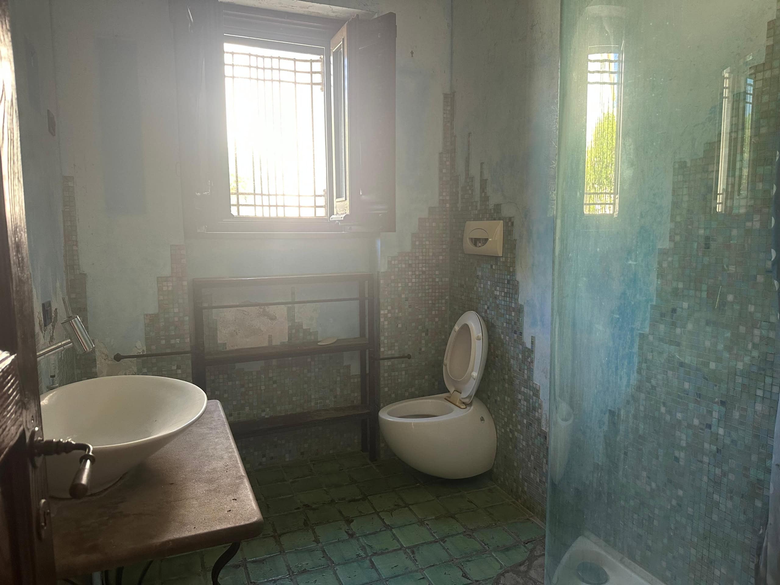 Interno di un bagno in fase di ristrutturazione con pareti in mosaico e finestra con grate di sicurezza.