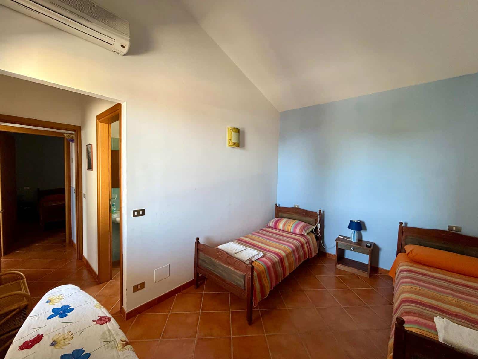 Camera che mostra una stanza con tre letti singoli, pareti bianche e azzurre, e un pavimento in piastrelle di terracotta, con una porta aperta che conduce a un'altra stanza.