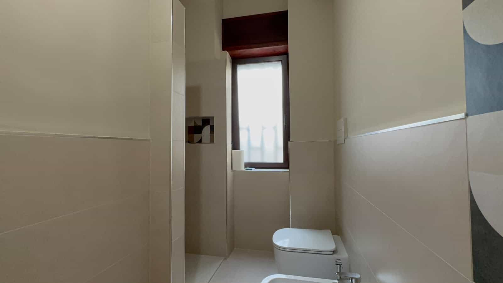 Interno di un bagno moderno con sanitari bianchi e pareti beige, finestra con tende leggere, in un appartamento in vendita a Milazzo.