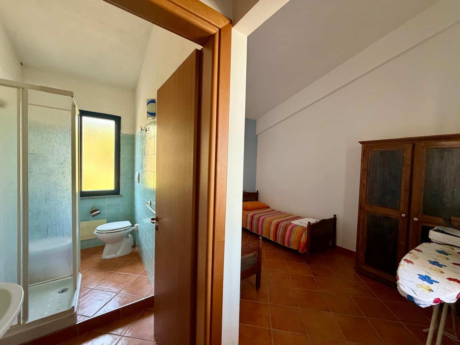 Interno di una casa con bagno e camera da letto, vista da una porta aperta, con arredi semplici e pavimento in piastrelle.