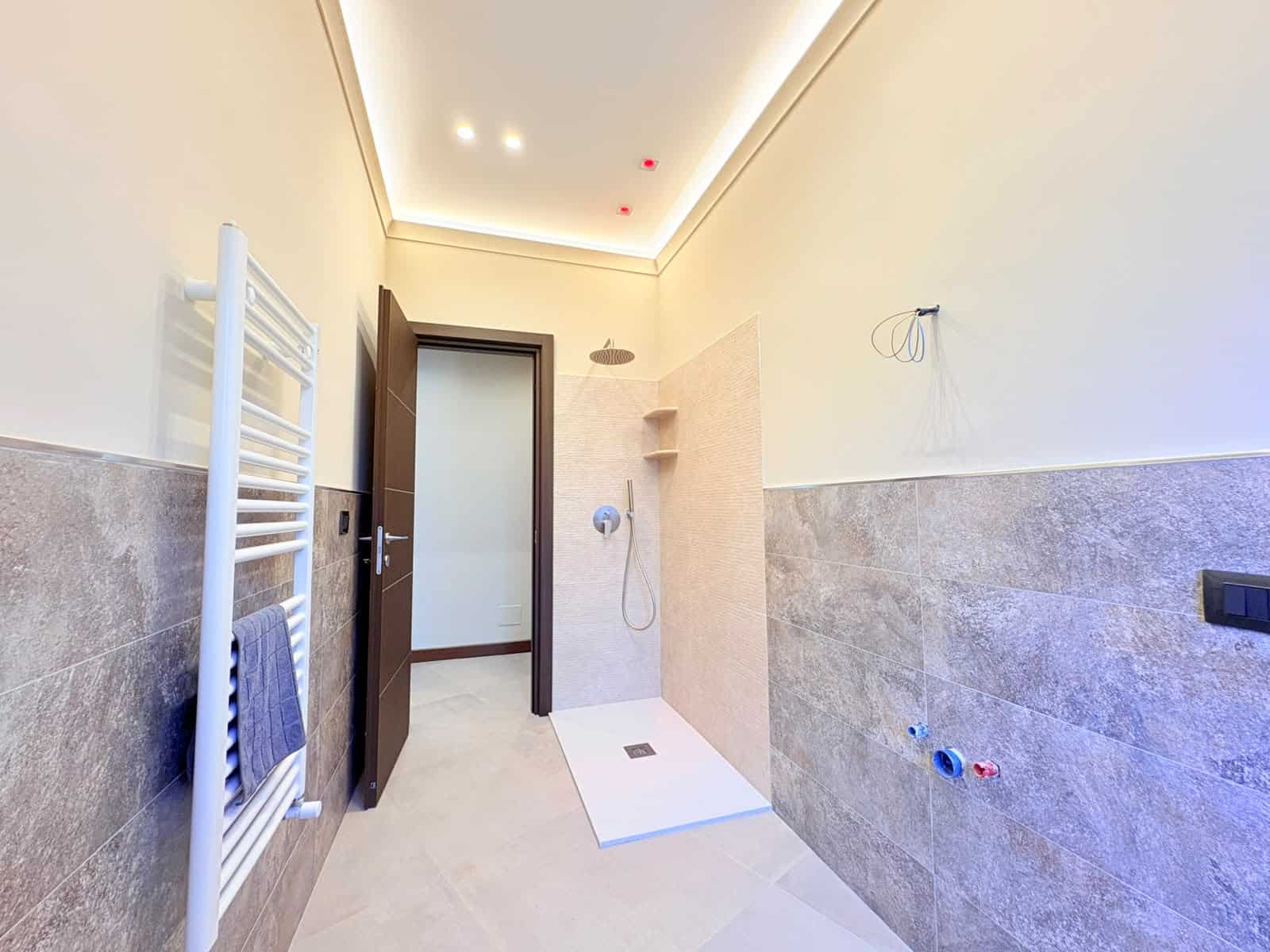 Bagno moderno con doccia a soffitto, pareti beige e piastrelle grigie, illuminato da luci a soffitto.