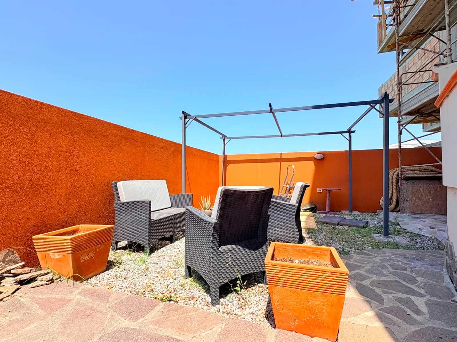 Spazio esterno con mobili da giardino, sedie e tavolo, in un cortile soleggiato con pareti arancioni e pavimentazione in pietra, vicino a un edificio in costruzione.