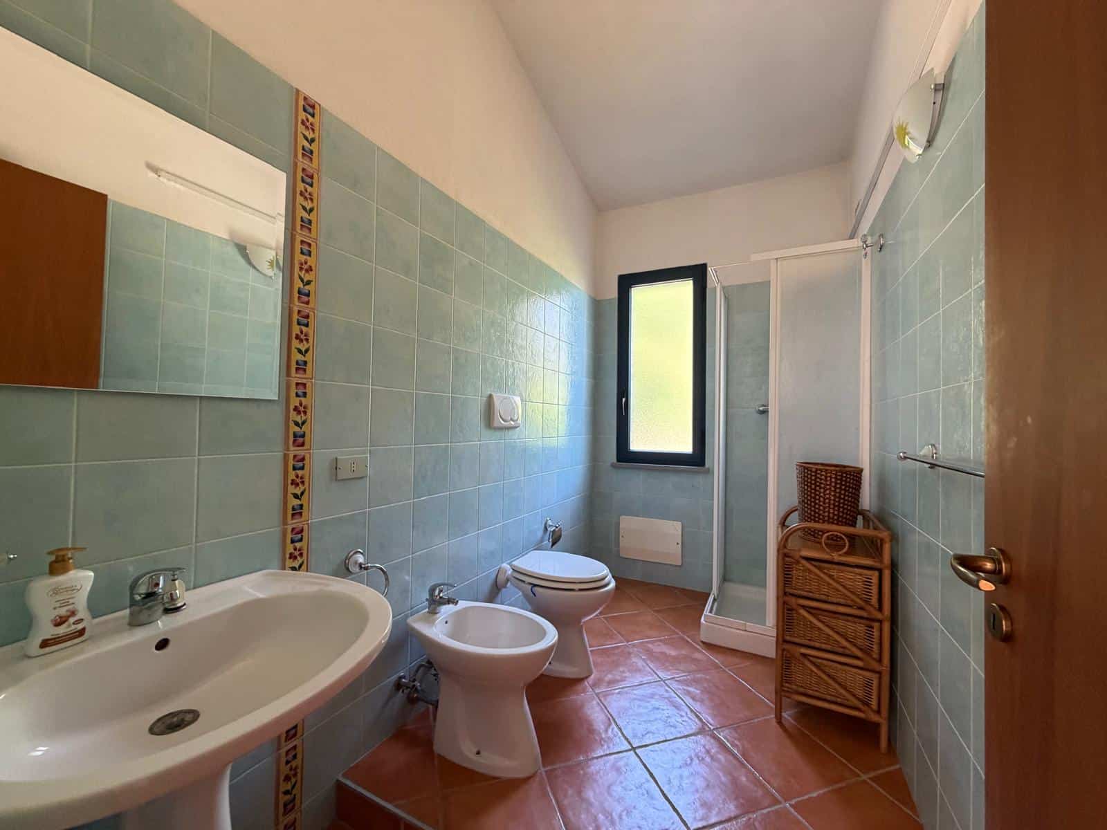Bagno spazioso con lavabo, bidet, WC e doccia, pareti rivestite di piastrelle azzurre e pavimento in terracotta, finestra per ventilazione naturale.