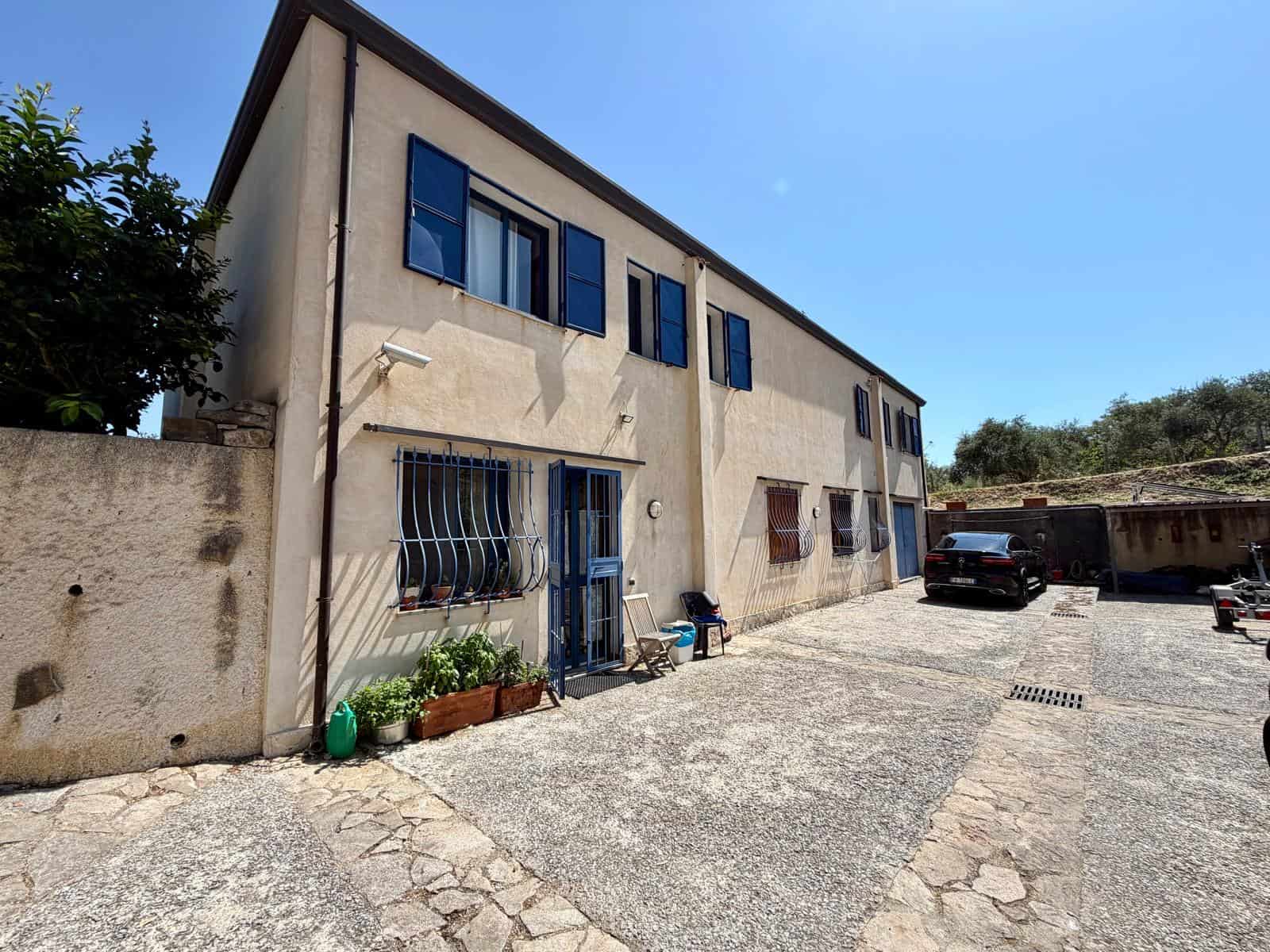 Edificio residenziale con facciata beige, finestre con persiane blu e ingresso principale, situato in un'area con parcheggio e vegetazione circostante.