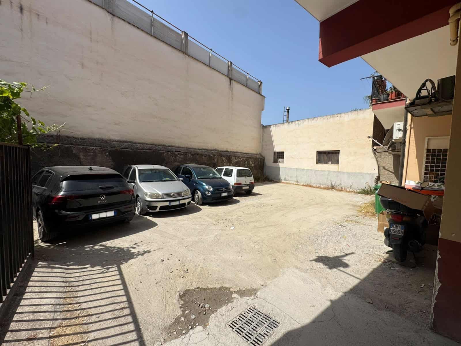 Parcheggio esterno con cinque auto e spazio libero, vicino a un edificio con pareti bianche e un muro alto.