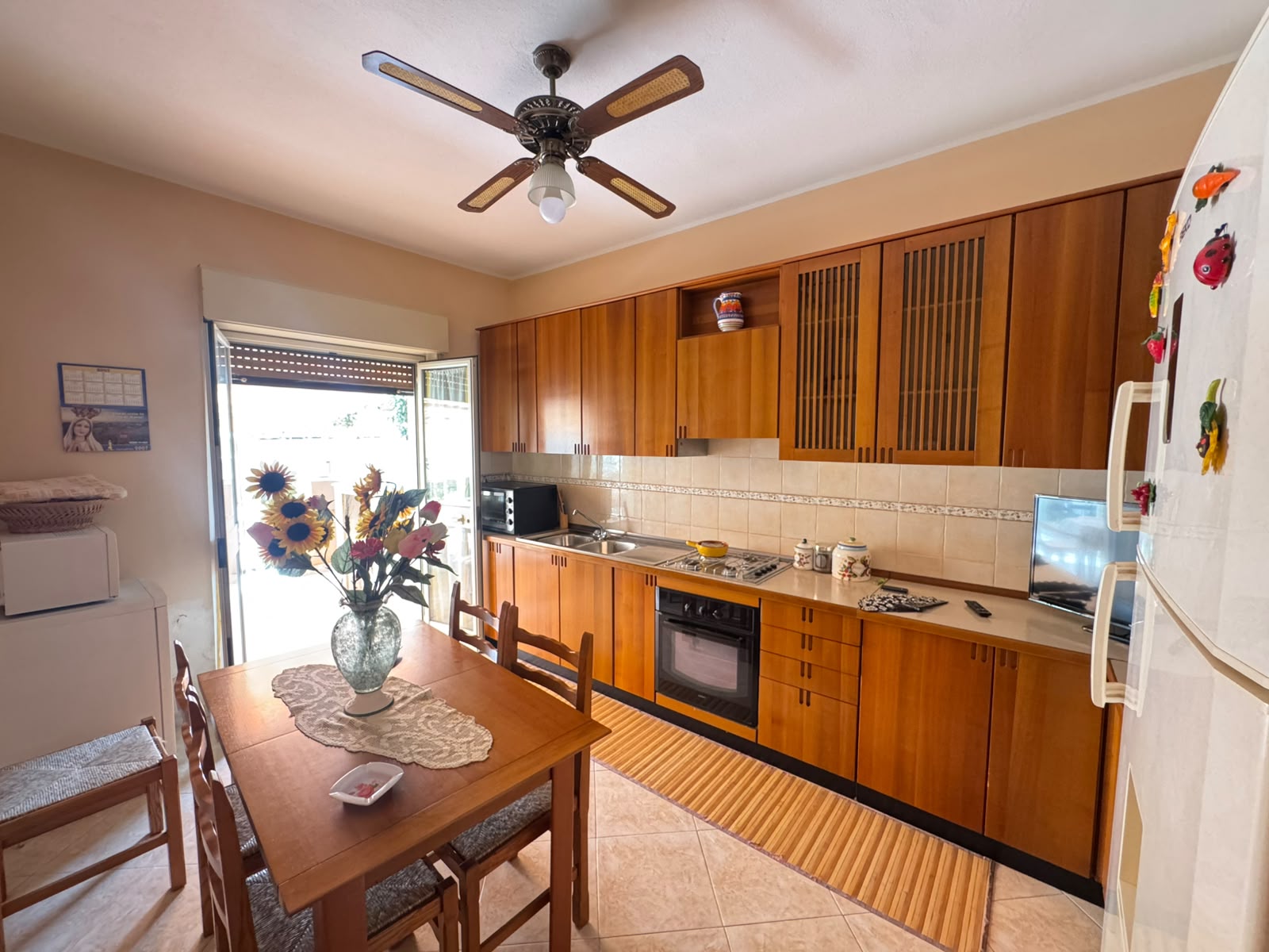 Cucina spaziosa con mobili in legno, tavolo con fiori e accessori moderni in un appartamento in vendita a Oliveri, Me.