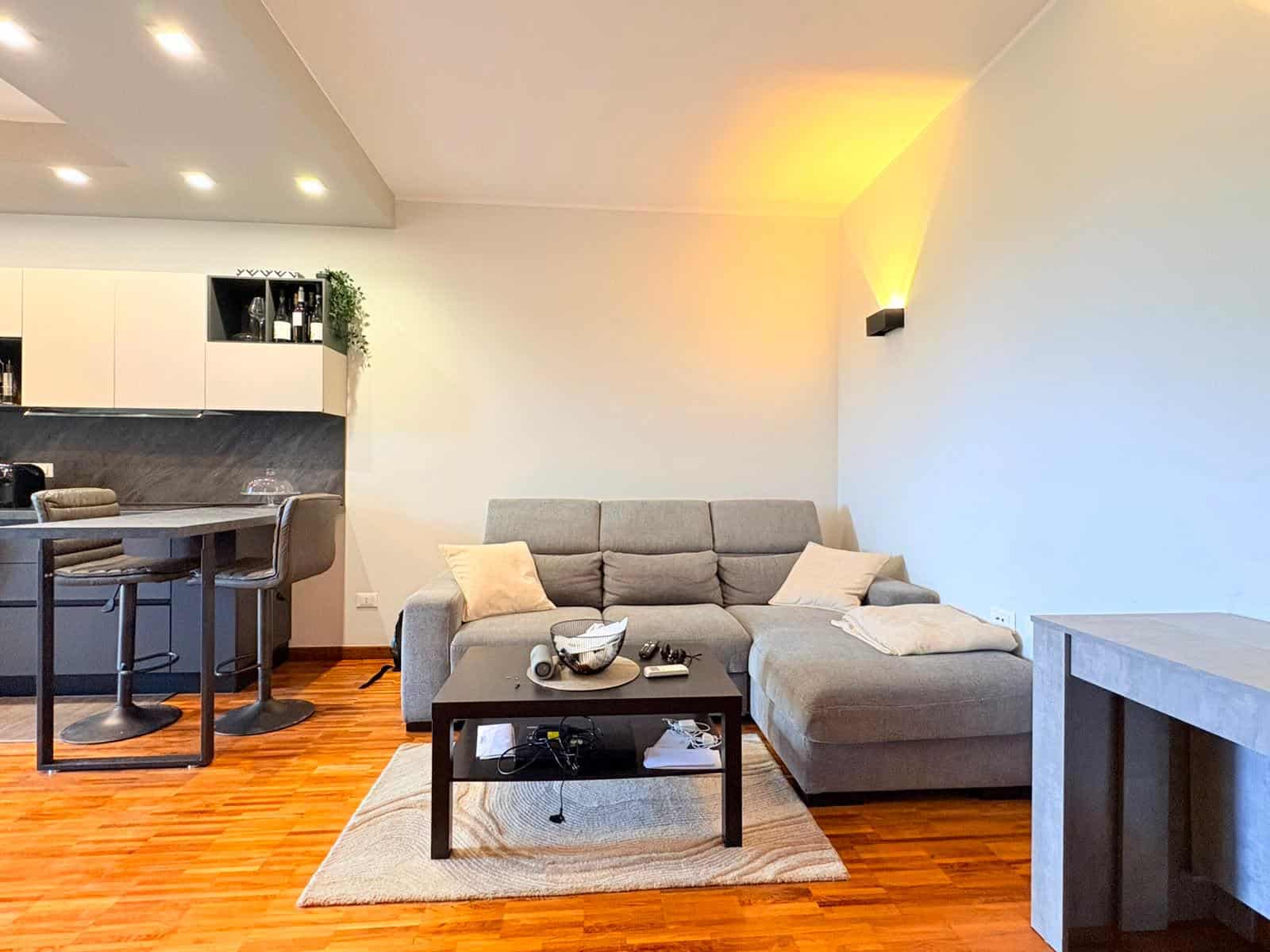 Interno di un appartamento moderno con soggiorno e cucina a vista, divano grigio e pavimento in parquet.