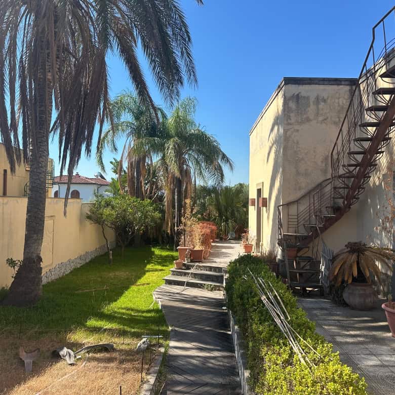 Giardino con palme e piante, vista su edificio con scala esterna in ferro, in zona residenziale a Milazzo.