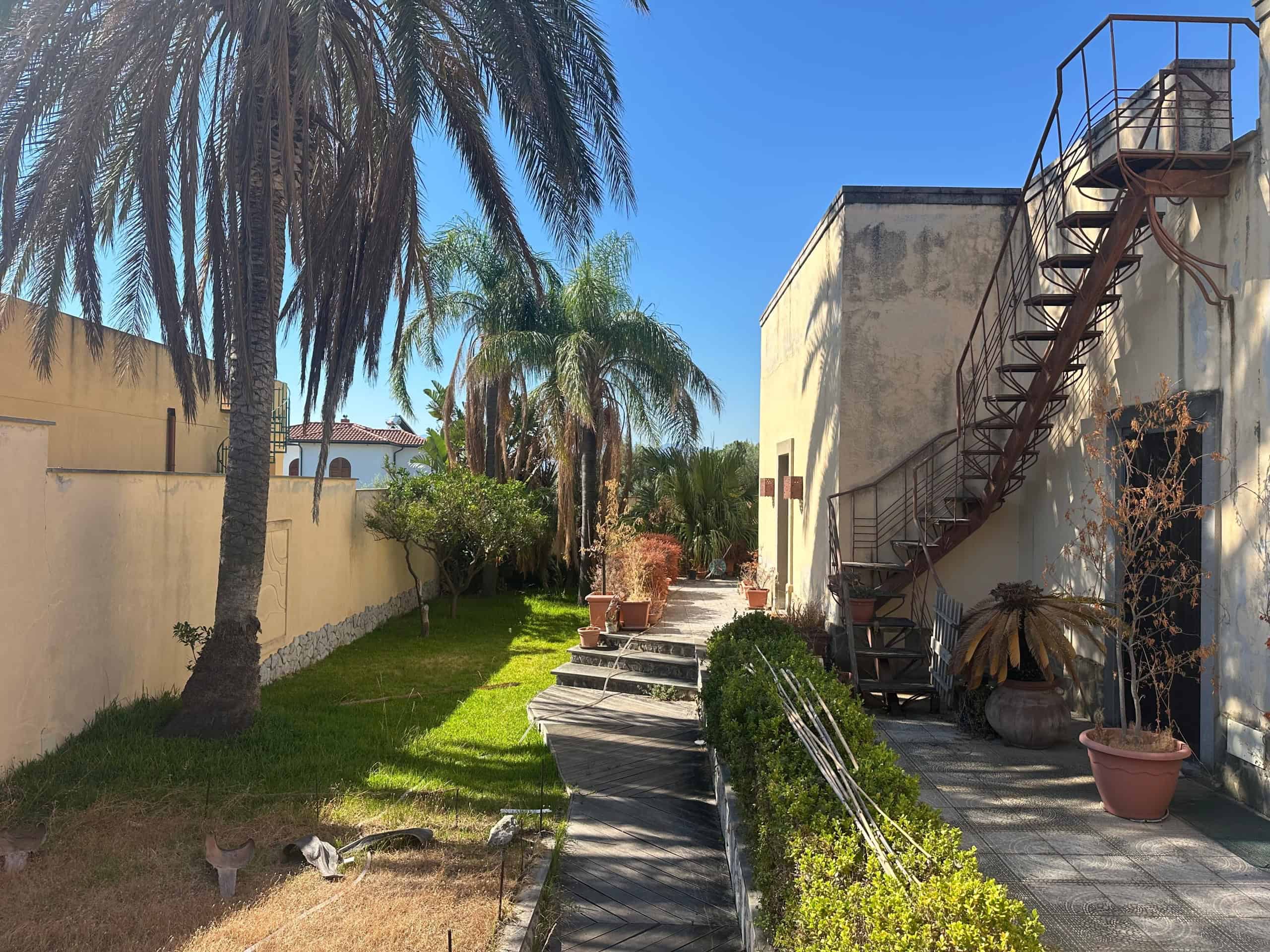 Giardino con palme e piante, vista su edificio con scala esterna in ferro, in zona residenziale a Milazzo.
