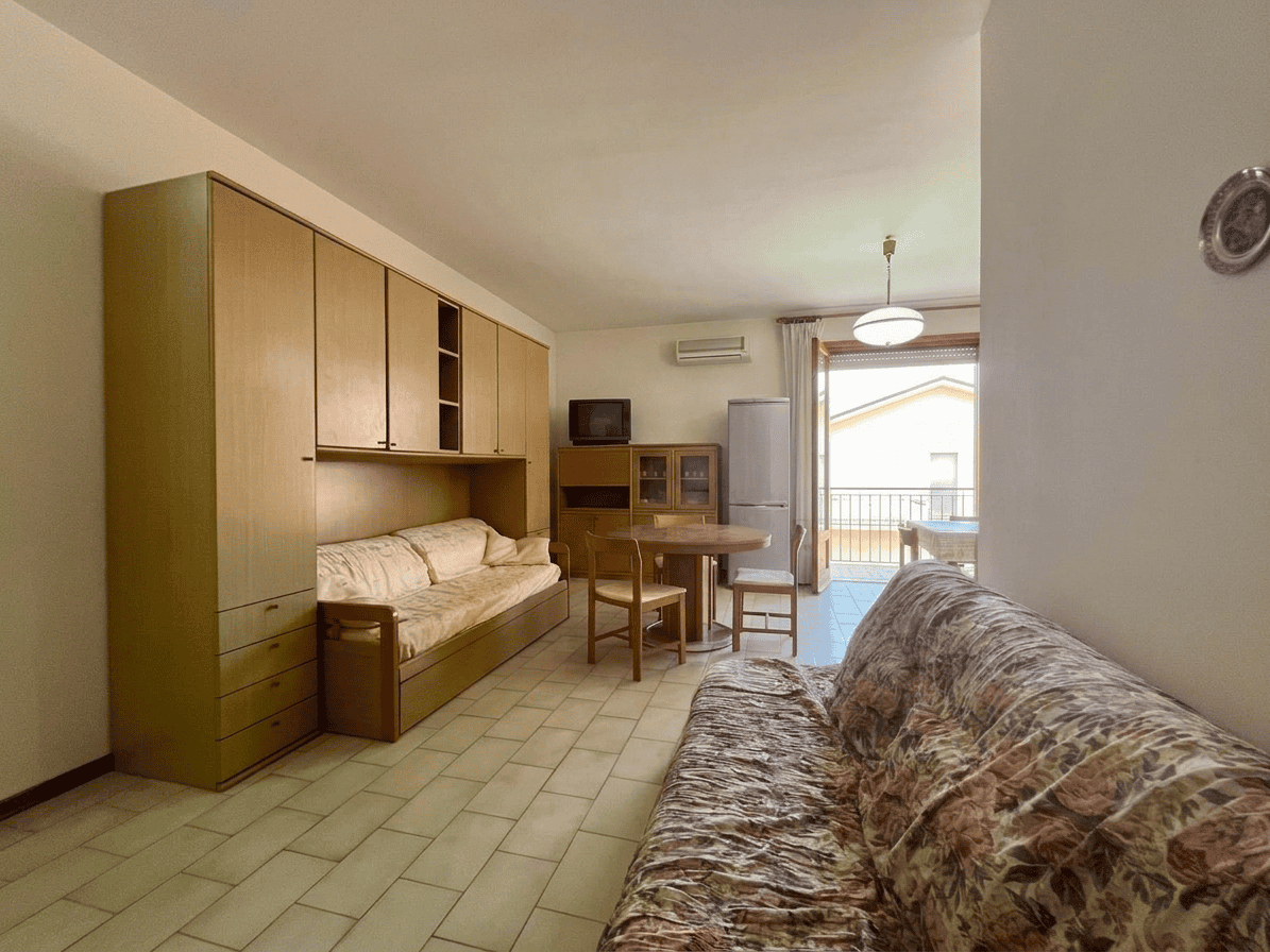 Interno di un appartamento con soggiorno arredato, divano, tavolo e cucina in vista, con balcone e vista sul mare.