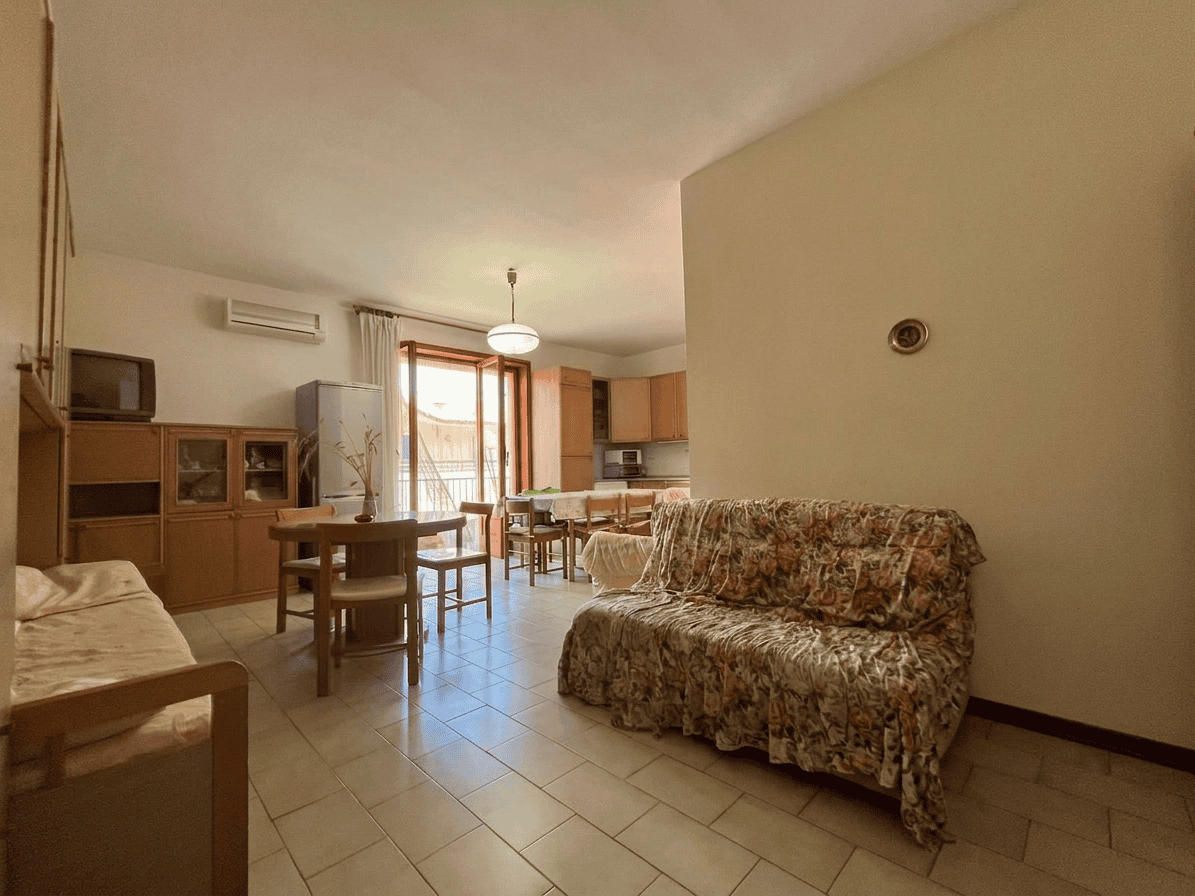 Interno di un appartamento con soggiorno e zona pranzo, arredato con divano, tavolo e sedie, con accesso a un balcone.