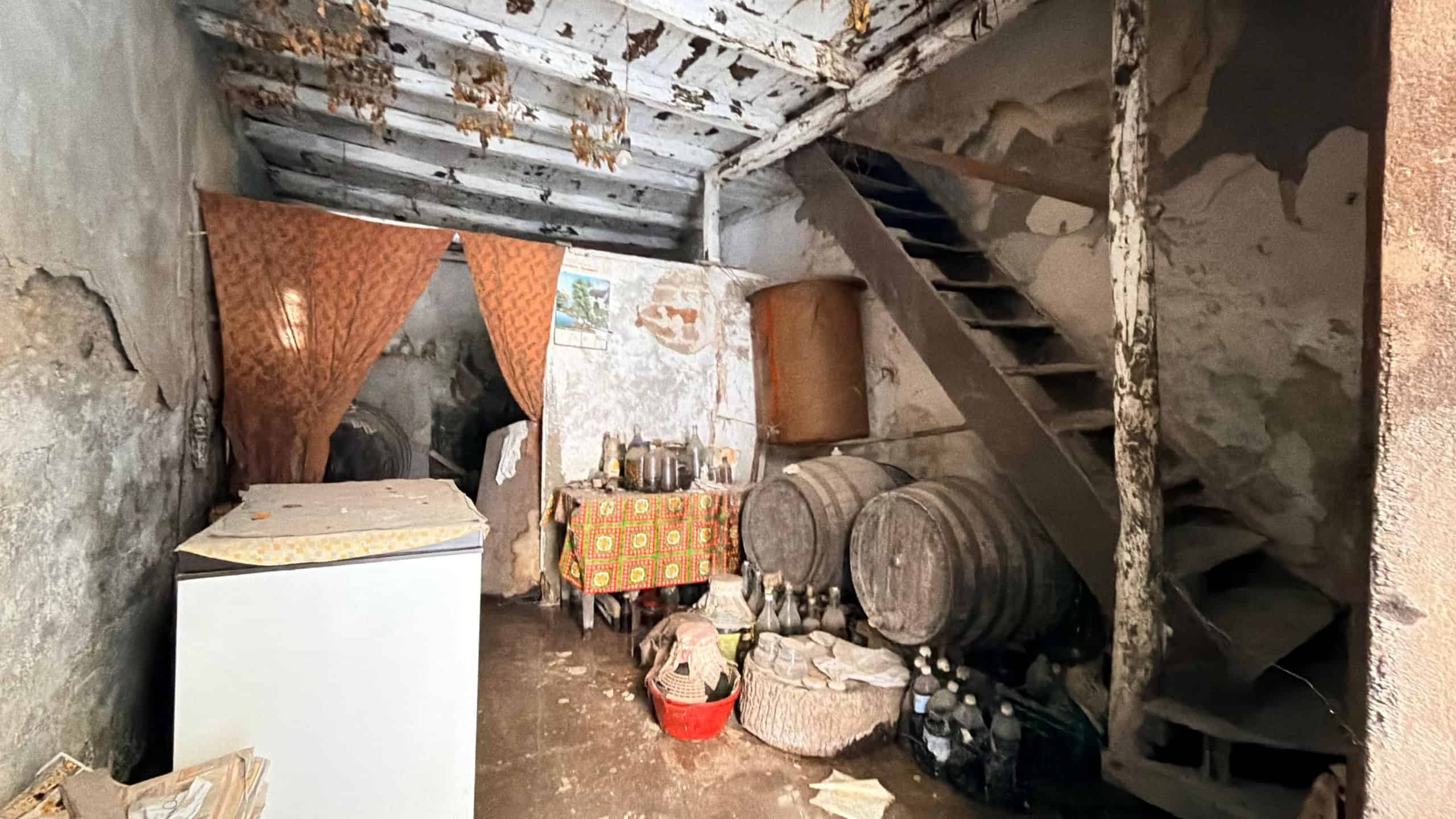 Interno di una cantina o soffitta trascurata con barili di vino, scatole e oggetti vari, pareti e soffitto in cattive condizioni.