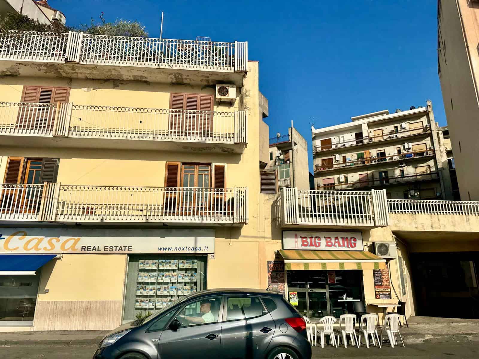 Edificio residenziale con negozi al piano terra e auto parcheggiata davanti, cielo sereno.
